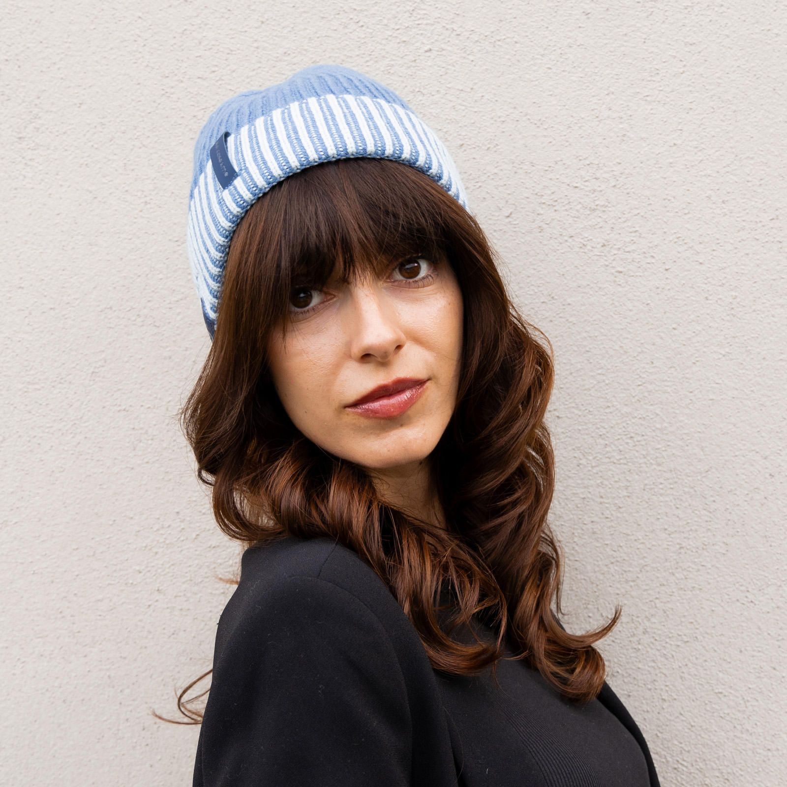 Una donna con lunghi capelli castani indossa un berretto azzurro con righe bianche contro una parete chiara.