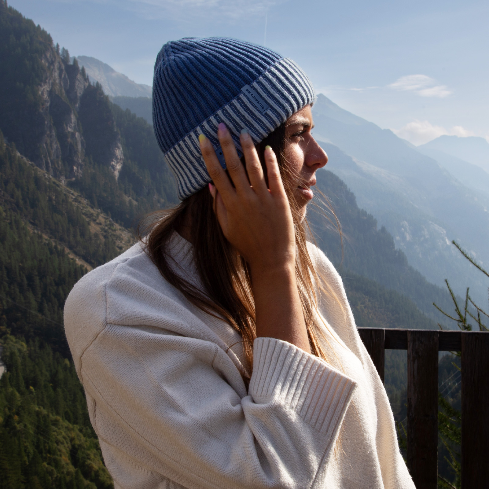 Donna con maglione bianco e berretto blu davanti a uno sfondo montano.