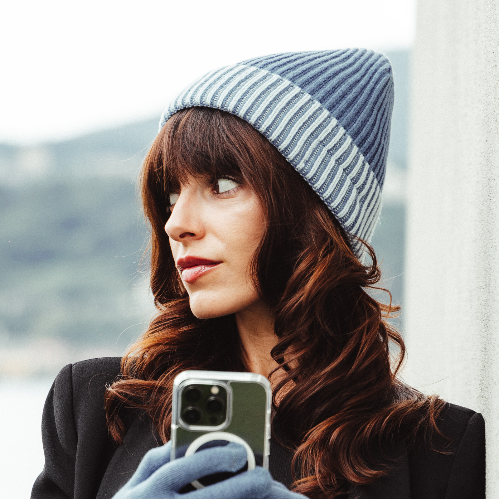 Una donna castana con berretto blu e cappotto tiene uno smartphone all'aperto.