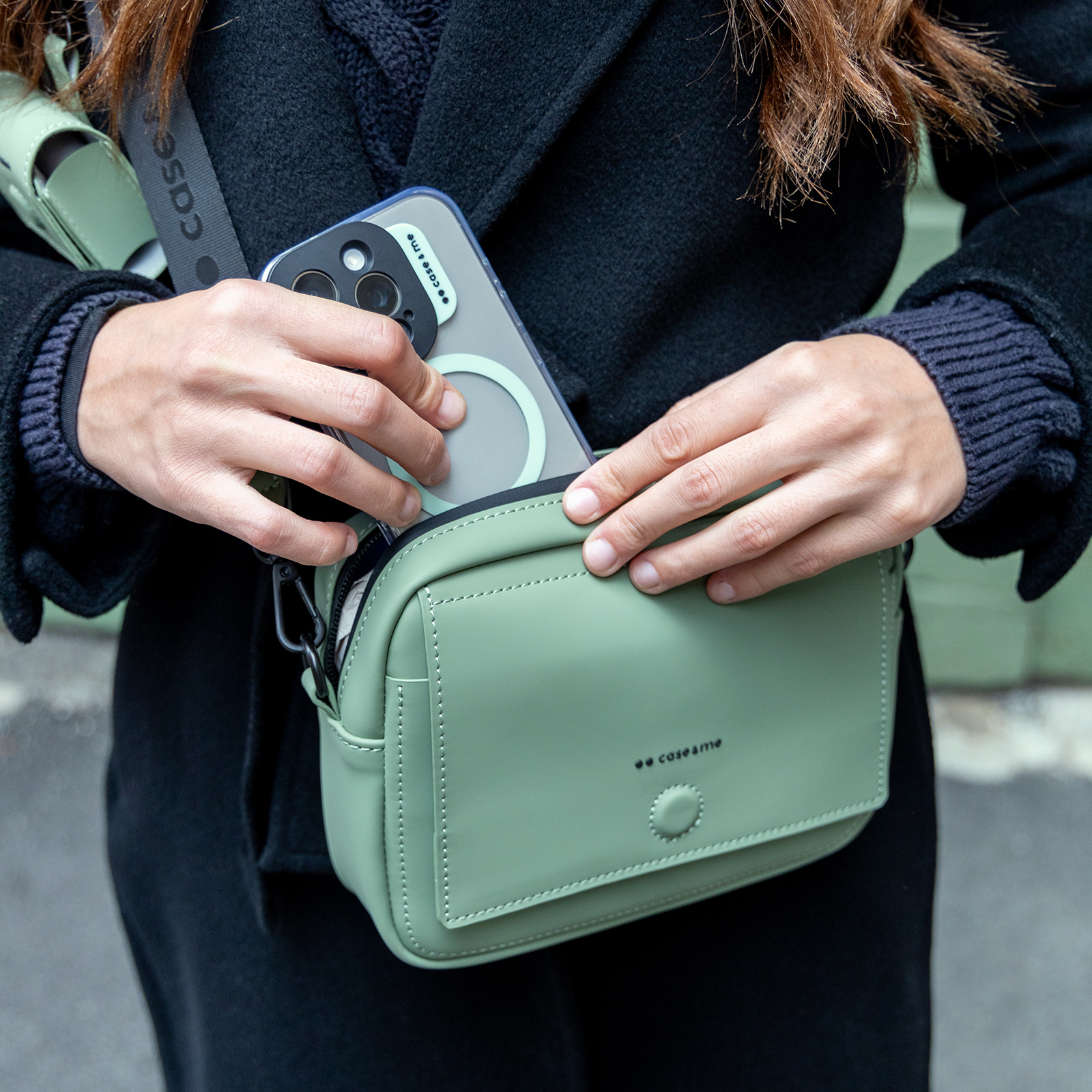 Persona con abrigo negro guarda un teléfono en un pequeño bolso bandolera verde.