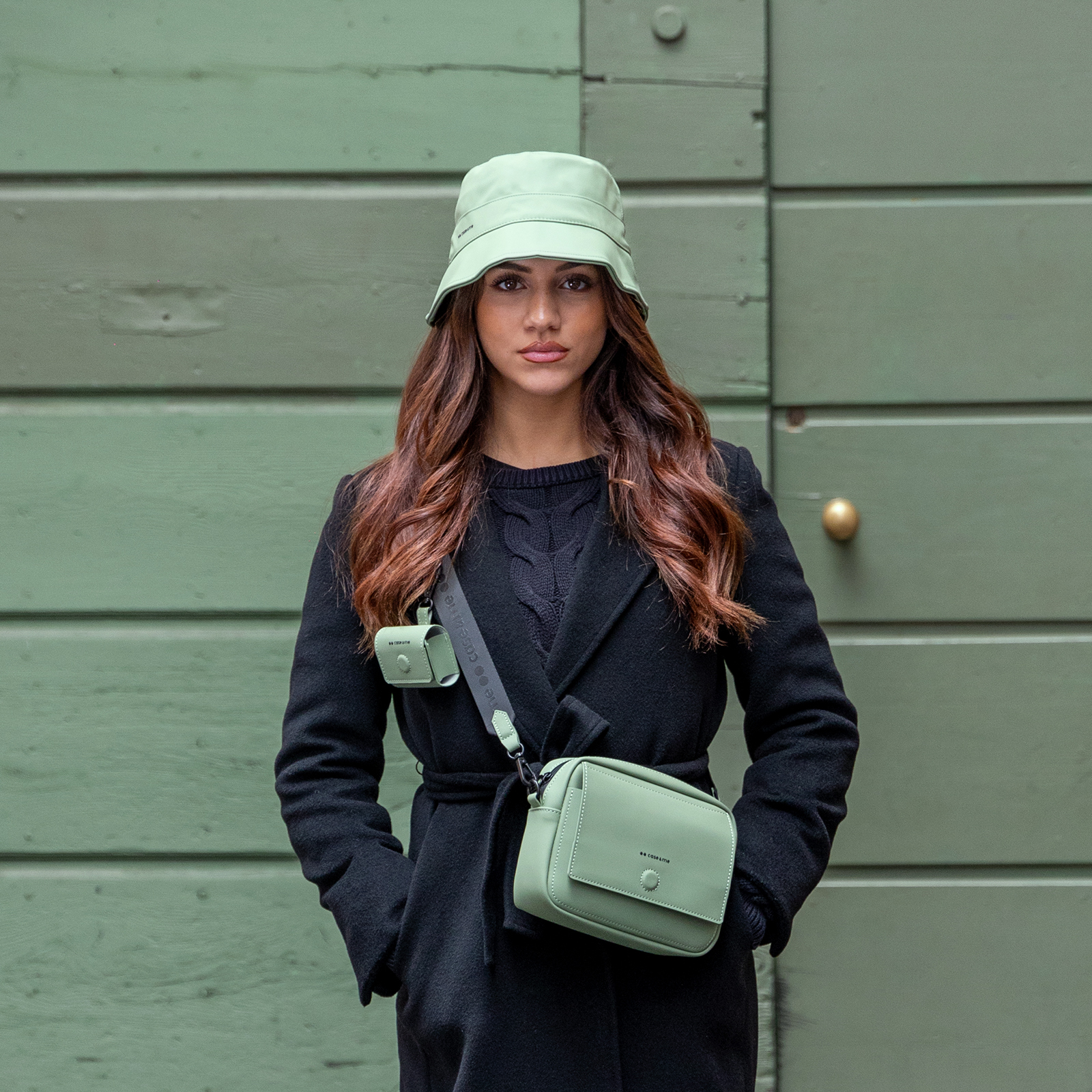 Una mujer con abrigo negro y sombrero de pescador verde lleva un bolso cruzado verde ante una puerta de madera verde.