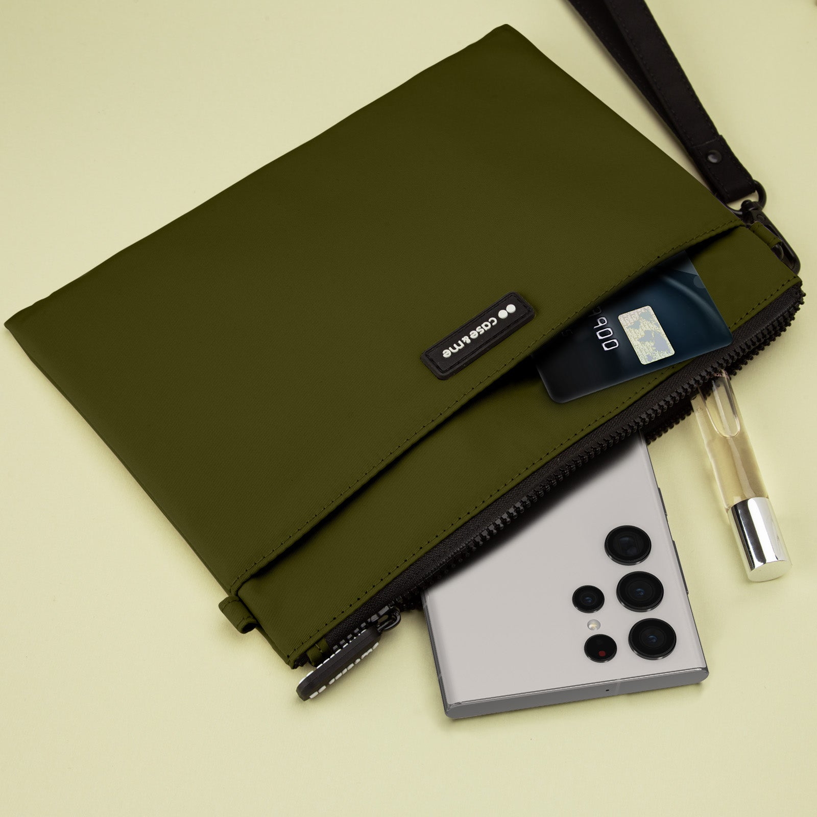Bolsa verde oliva con un teléfono inteligente, tarjeta de crédito y frasco de perfume sobre fondo claro.