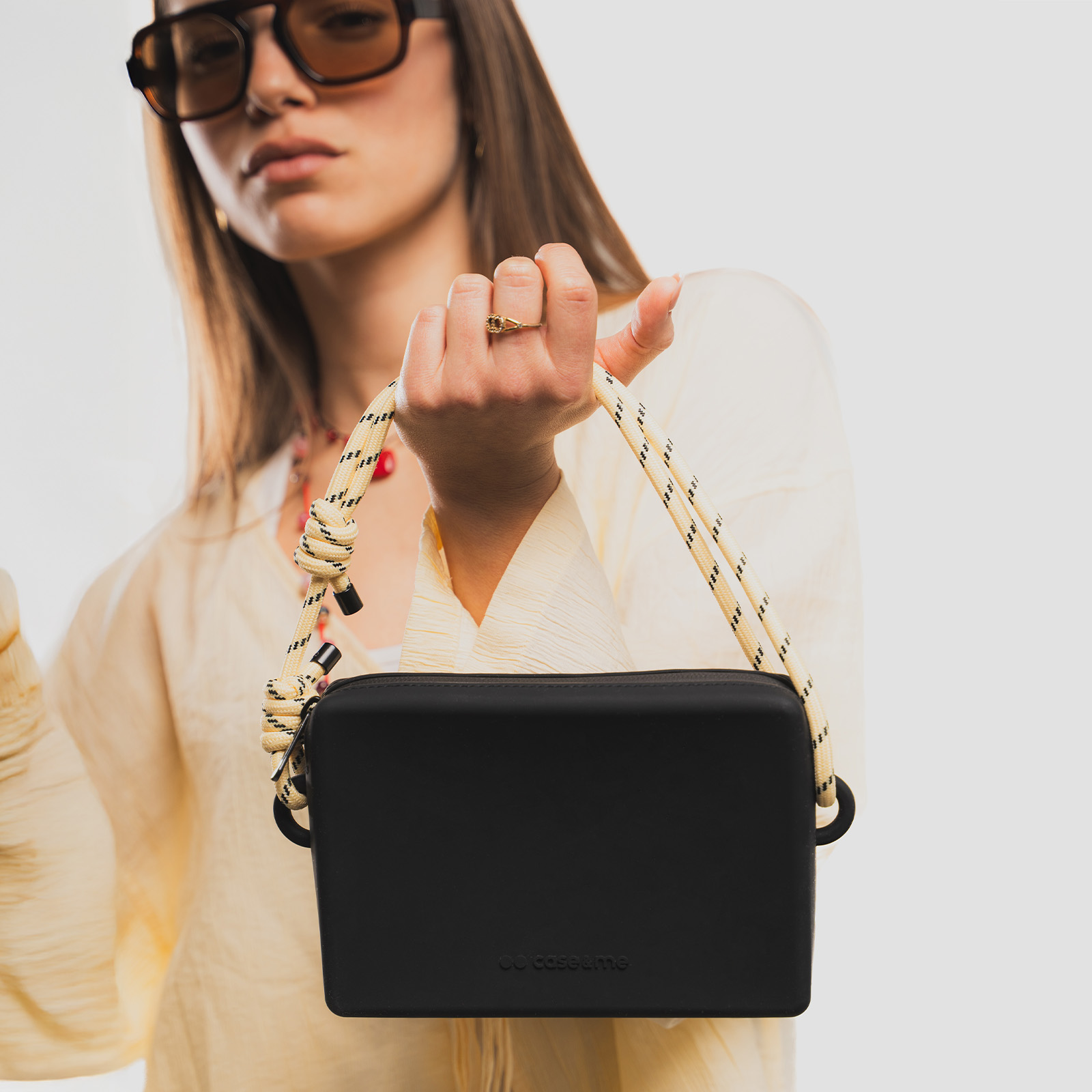 Una mujer con gafas de sol sostiene un bolso bandolera negro con correa de cuerda beis sobre fondo neutro.