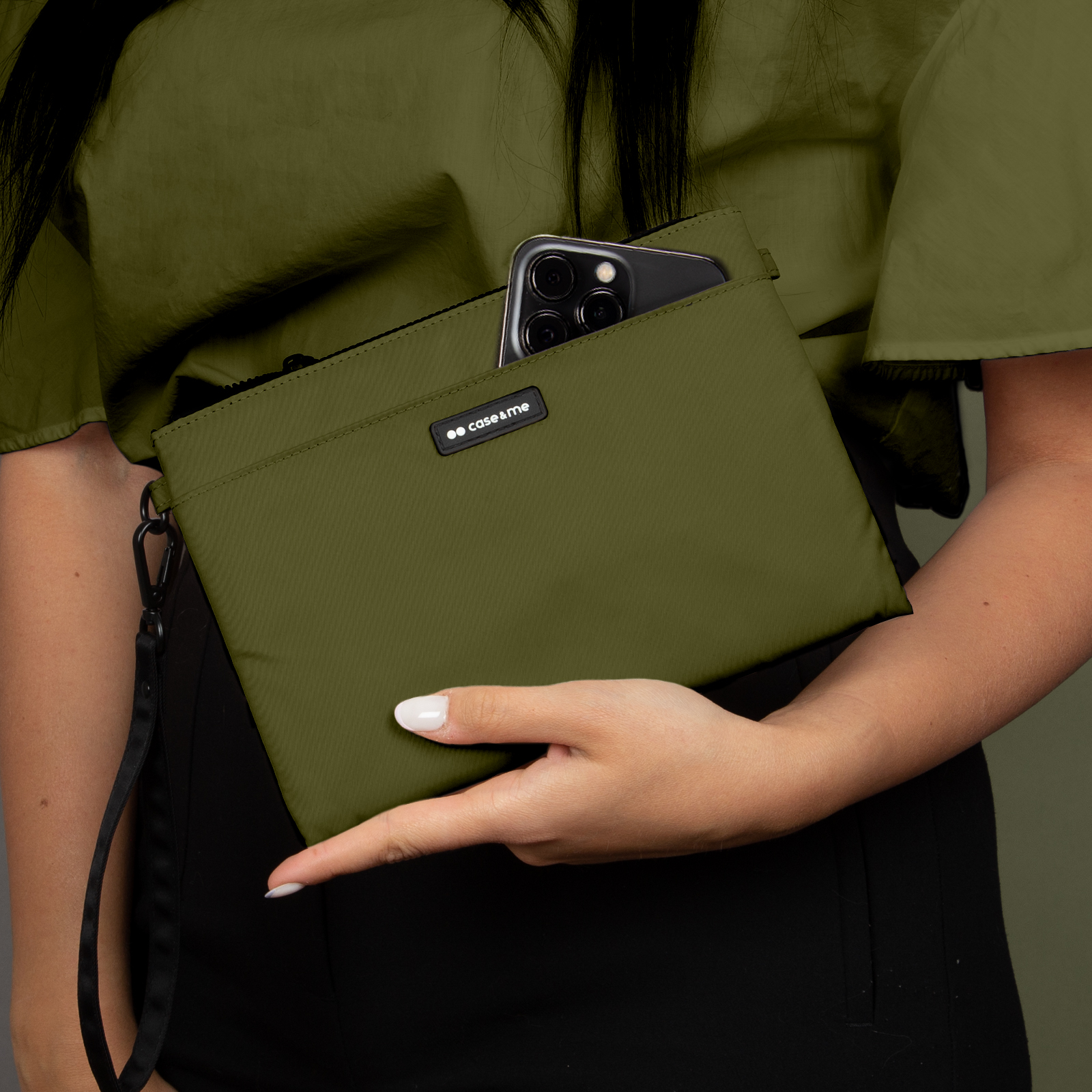 Una persona sostiene un bolso verde oliva con un smartphone, vistiendo una camisa verde y pantalones negros.