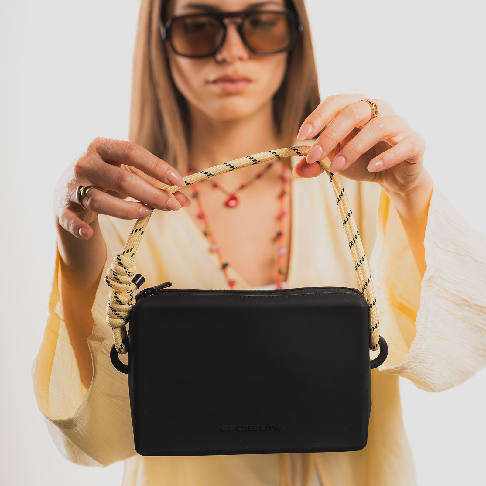 Una mujer sostiene un bolso bandolera negro, rectangular y pequeño con correa de cuerda gruesa.