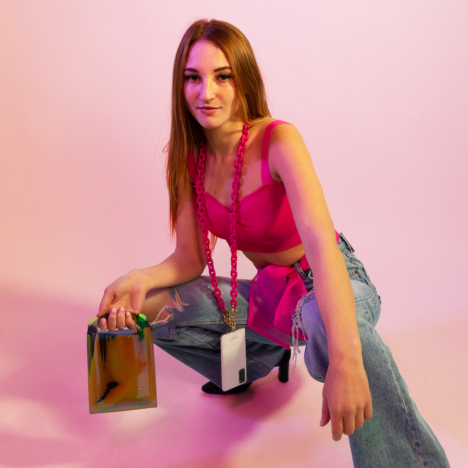 Una mujer joven con cabello largo y rojizo en cuclillas, vestida con top rosa y jeans, sostiene un bolso pequeño frente a fondo rosa.