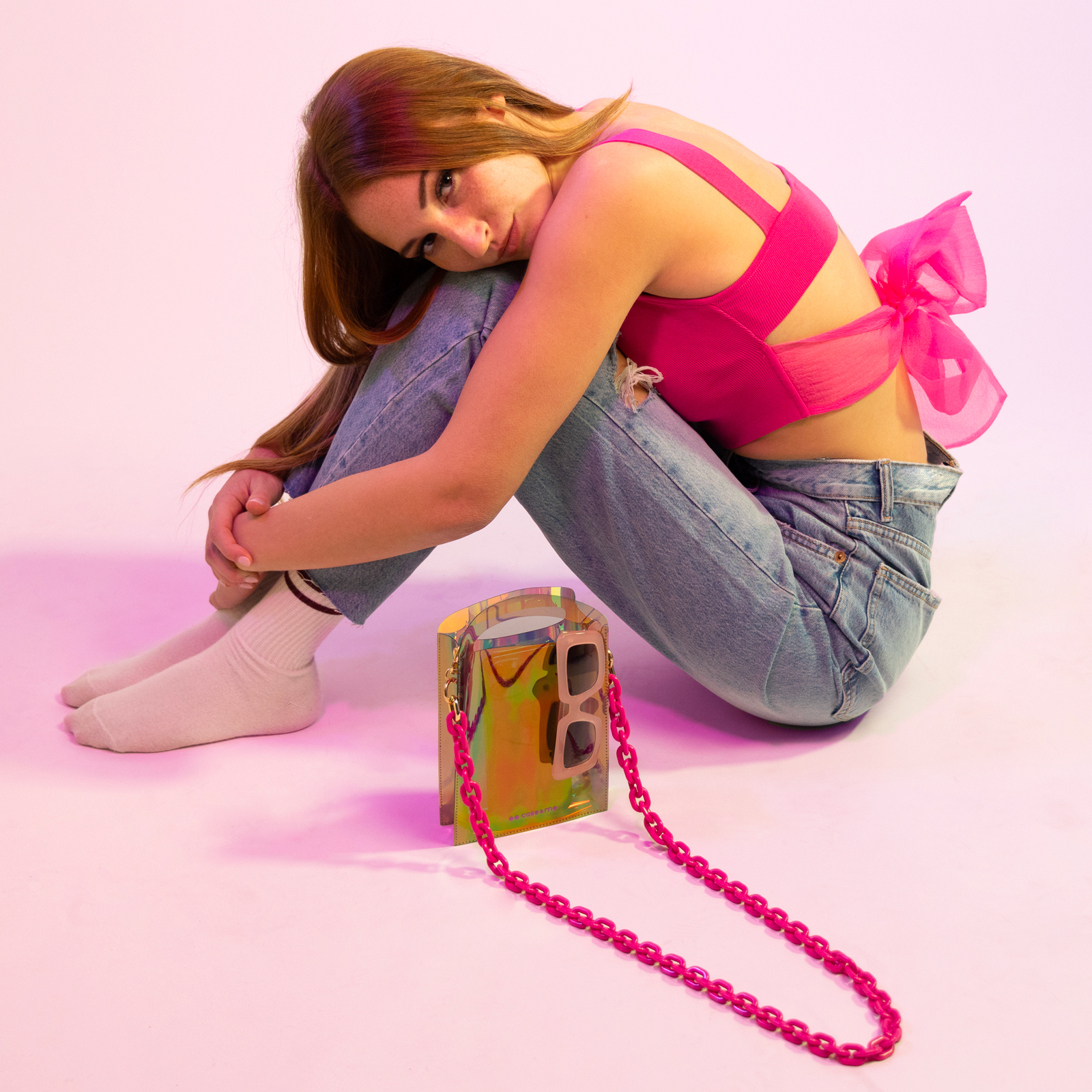 Mujer con jeans y top rosa con lazo sentada en el suelo con un bolso holográfico.