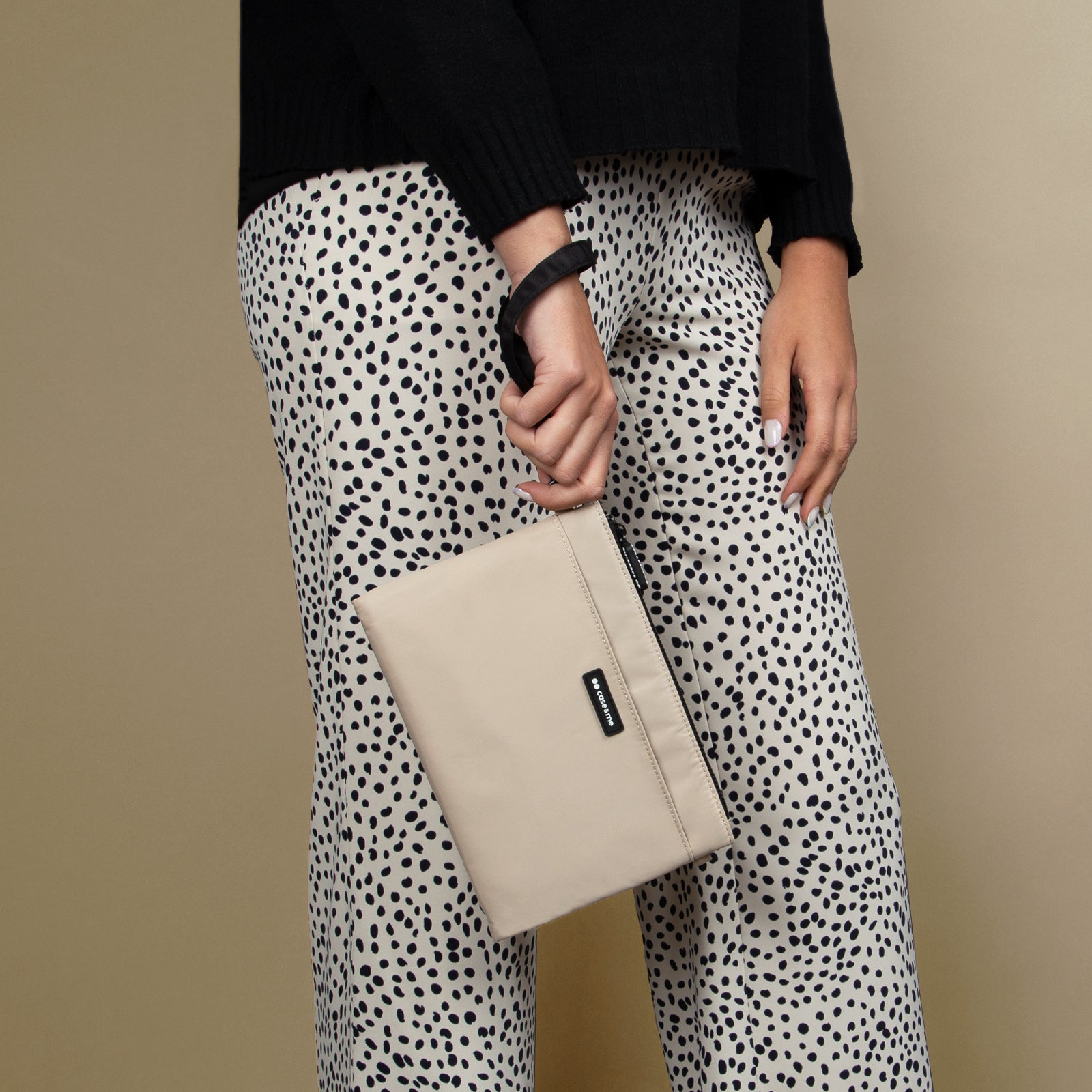 Una persona con un suéter negro y pantalones estampados en blanco y negro sostiene un pequeño bolso beige frente a ella.