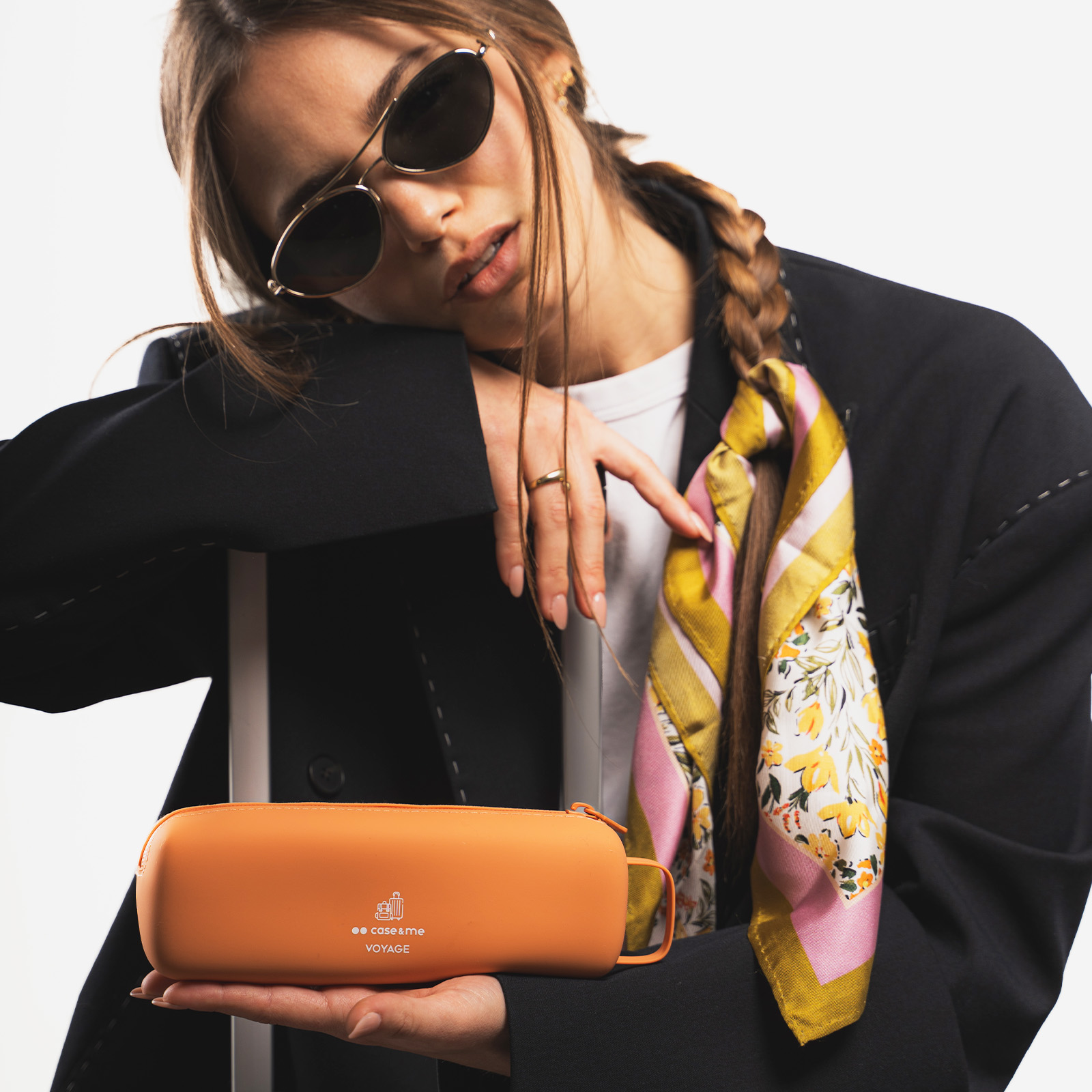 Una mujer elegante con gafas de sol y blazer sostiene un estuche naranja, posando sobre fondo blanco.