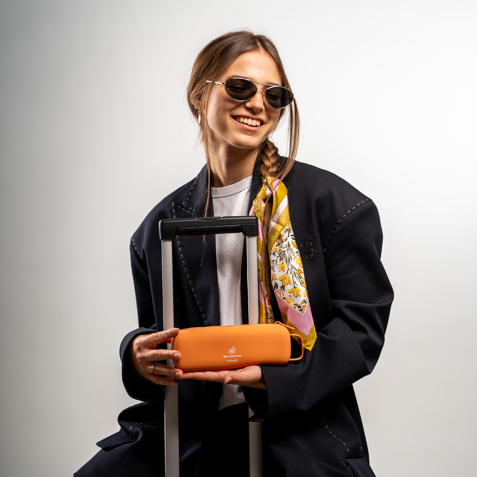 Una mujer sonriente con gafas de sol y blazer grande sostiene un estuche naranja frente al asa de una maleta.