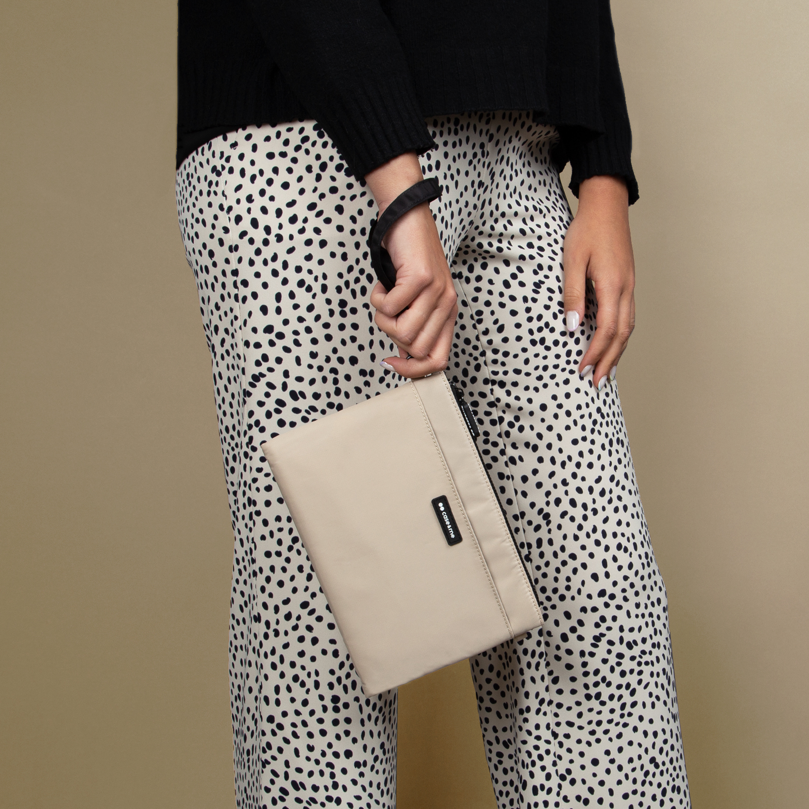 Una persona con un suéter negro y pantalones estampados sostiene un bolso beige pequeño.
