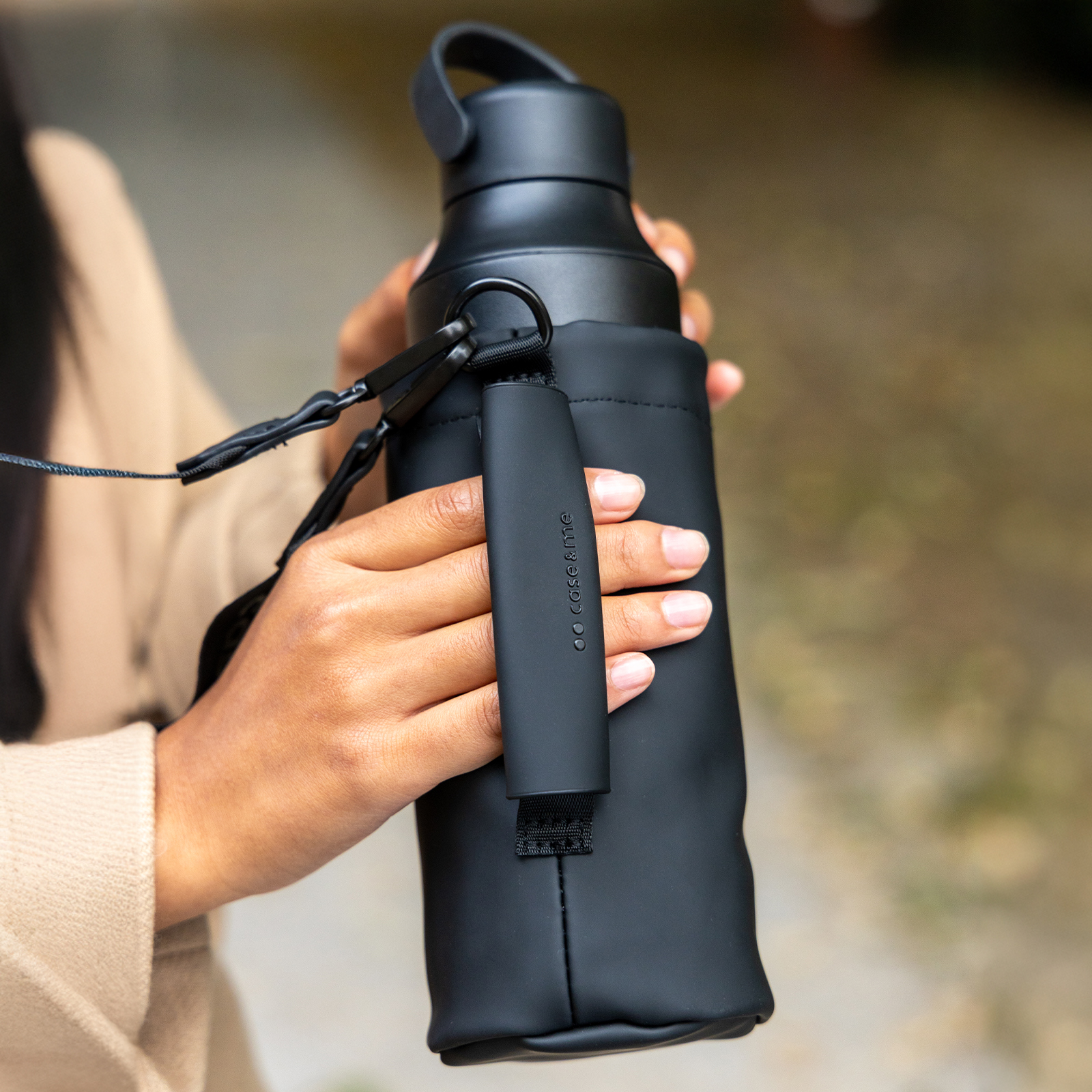 Una persona sostiene una botella de agua negra con correa y funda protectora al aire libre.