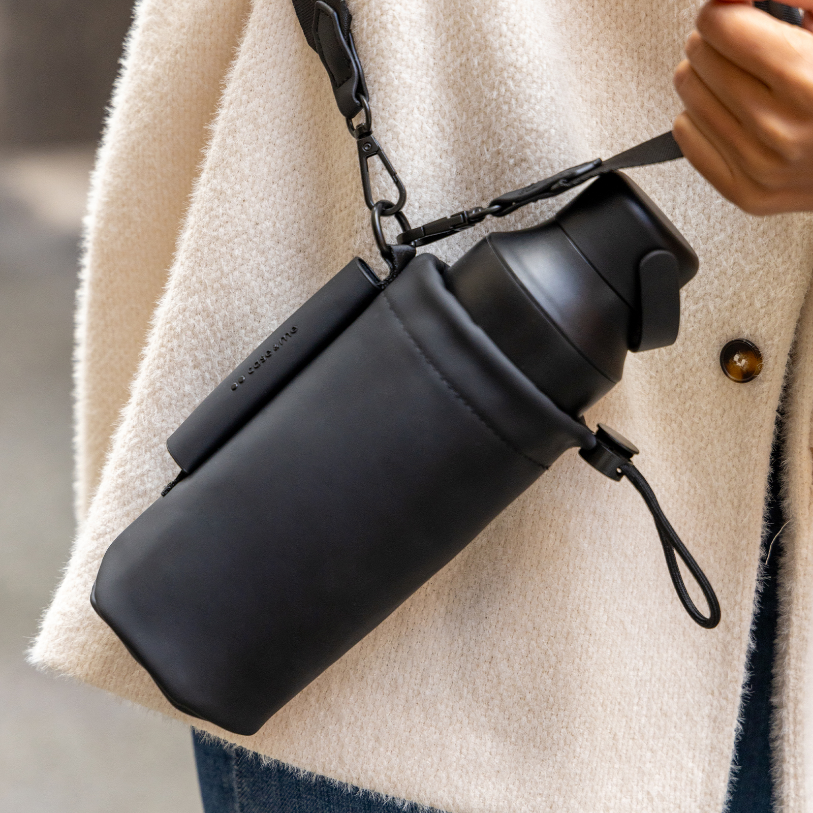 Una botella de agua negra en una funda negra con correa, llevada sobre un abrigo blanco.