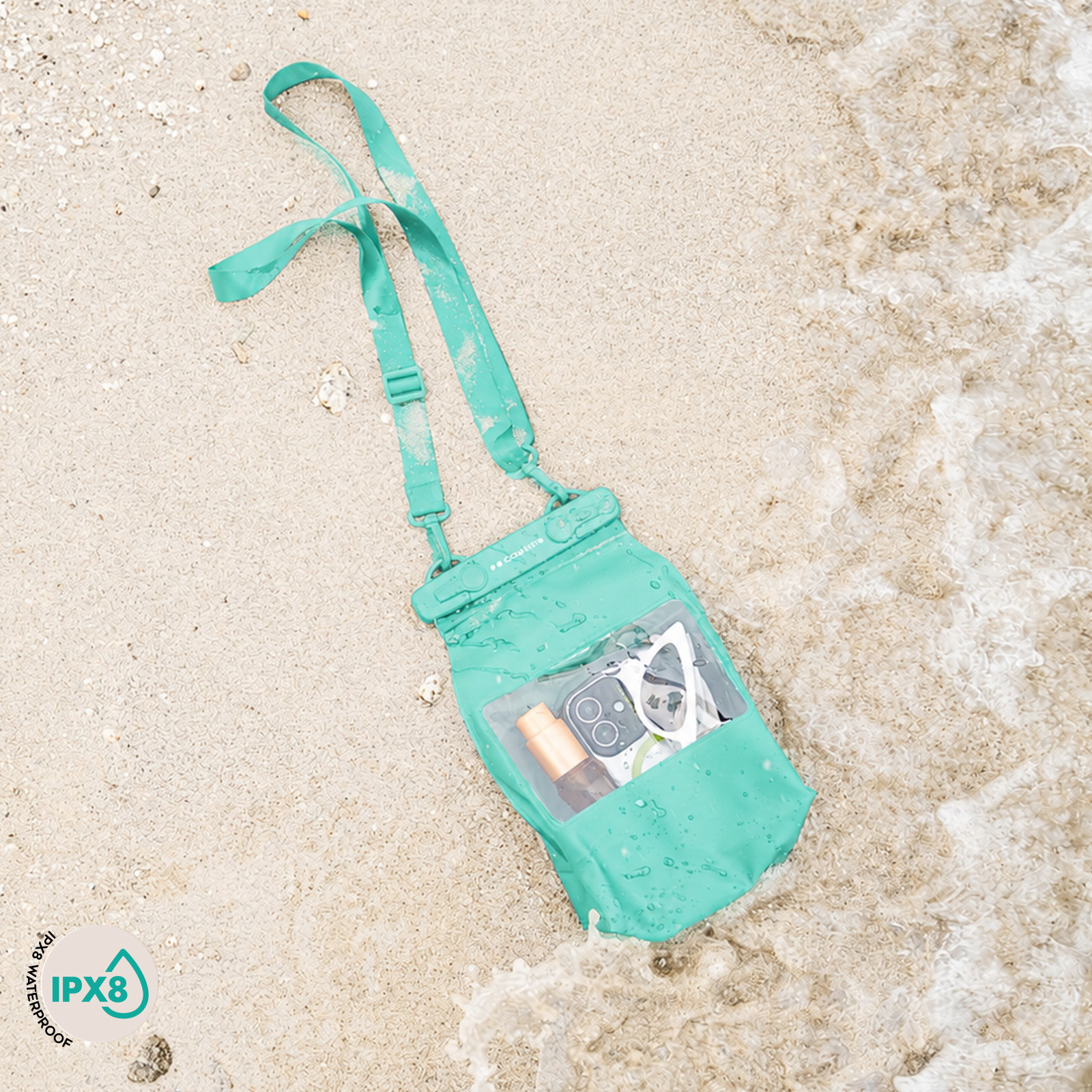 Una bolsa impermeable turquesa con teléfono y gafas de sol yace en la arena mojada cerca del mar.