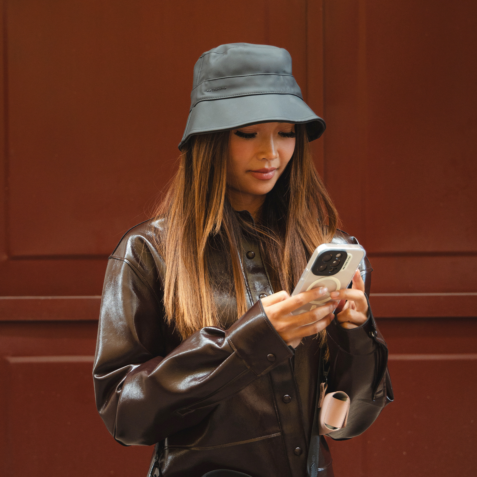 Mujer con sombrero de pescador oscuro y chaqueta de charol marrón mirando su smartphone ante fondo rojo.