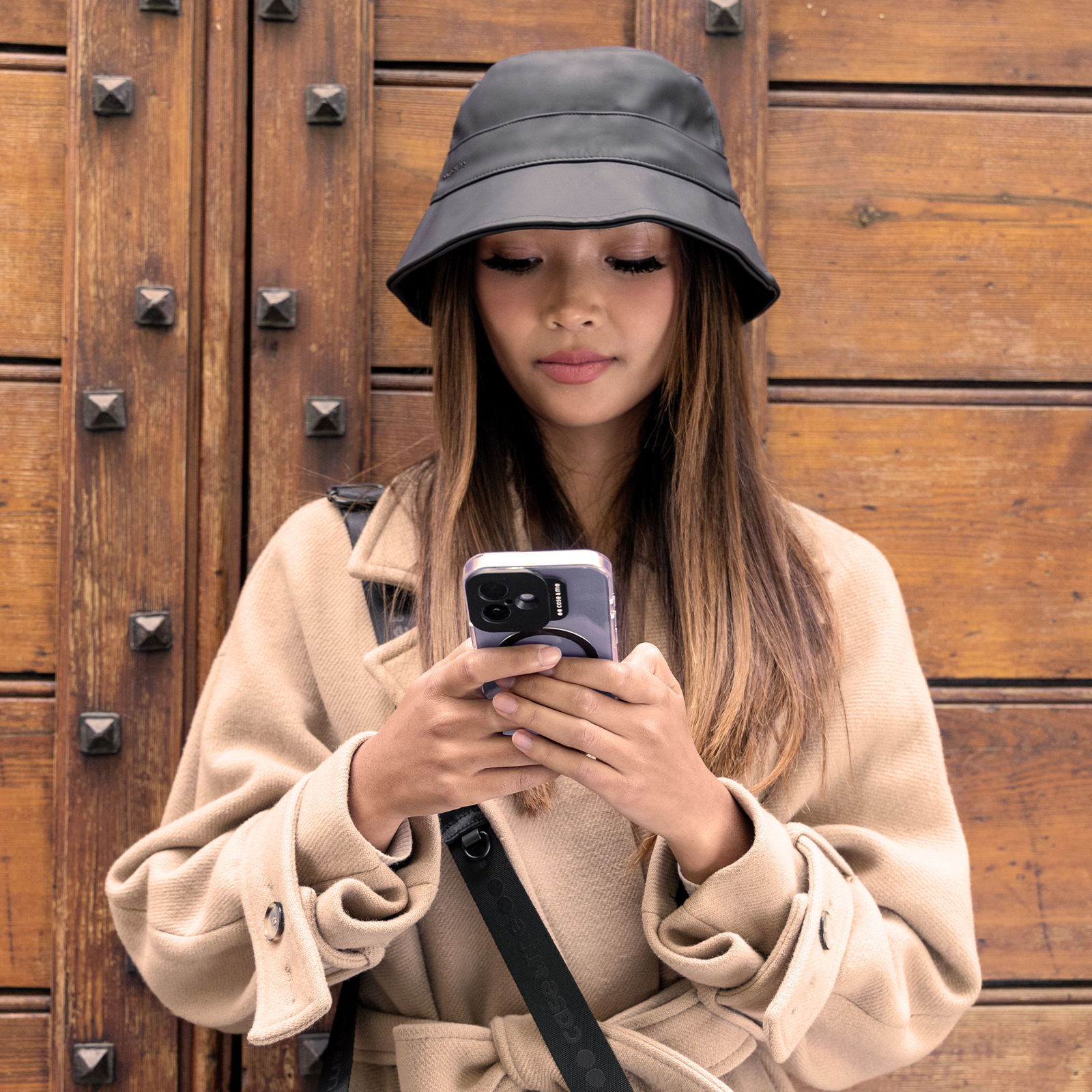 Mujer con sombrero negro y abrigo beige mirando su teléfono inteligente ante una puerta de madera.
