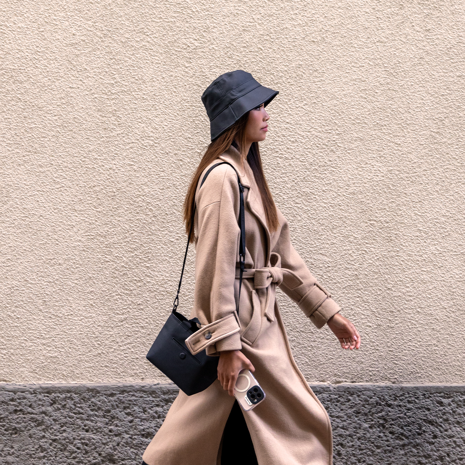 Una mujer con abrigo beige y sombrero negro camina junto a una pared beige.