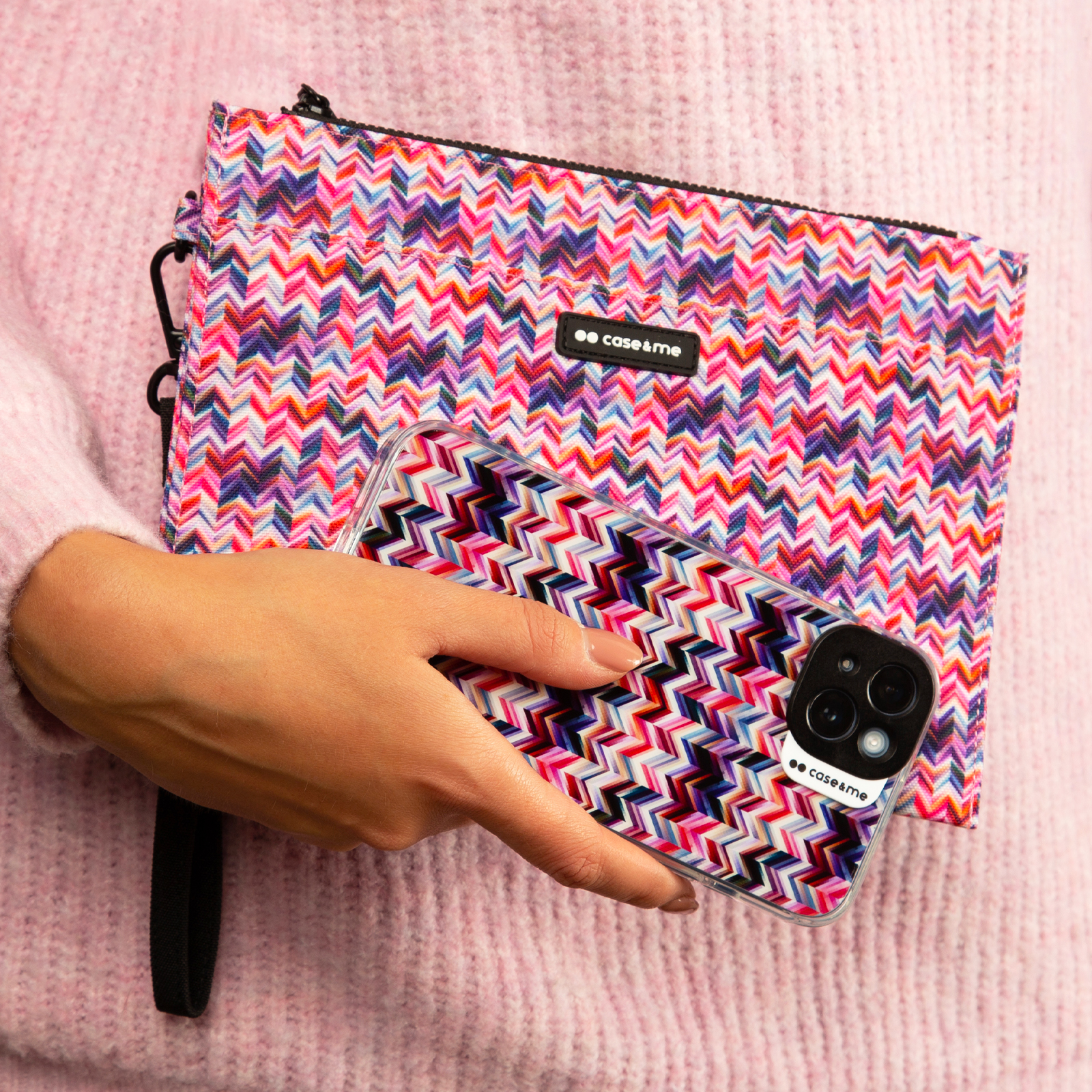 Una persona sostiene una funda de móvil y un estuche a juego con patrón de cheurón contra un suéter rosa.