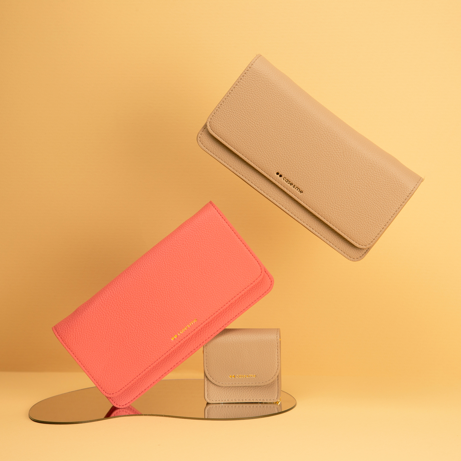 Tres carteras de cuero en coral y beige flotan sobre fondo amarillo con un pequeño espejo.