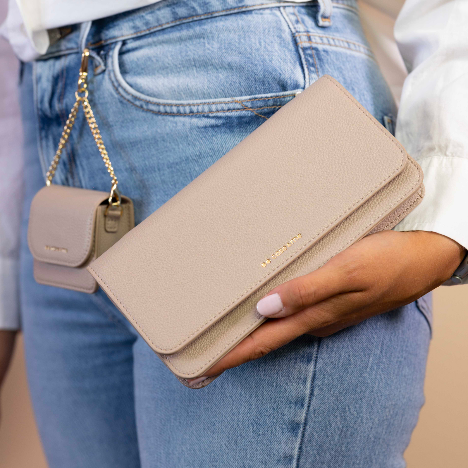 Una mano sostiene un bolso bandolera beige frente a unos vaqueros azules, con una pequeña bolsa colgando de una cadena.