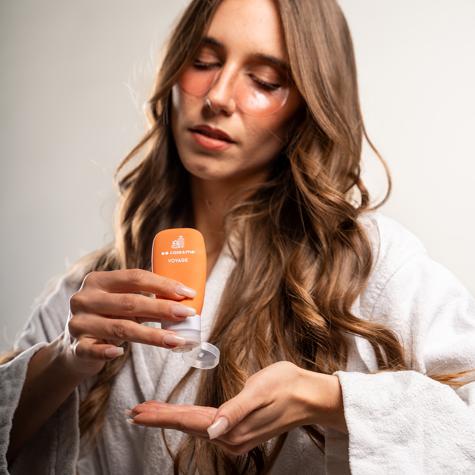 Una mujer con albornoz blanco se aplica crema facial desde un tubo naranja.