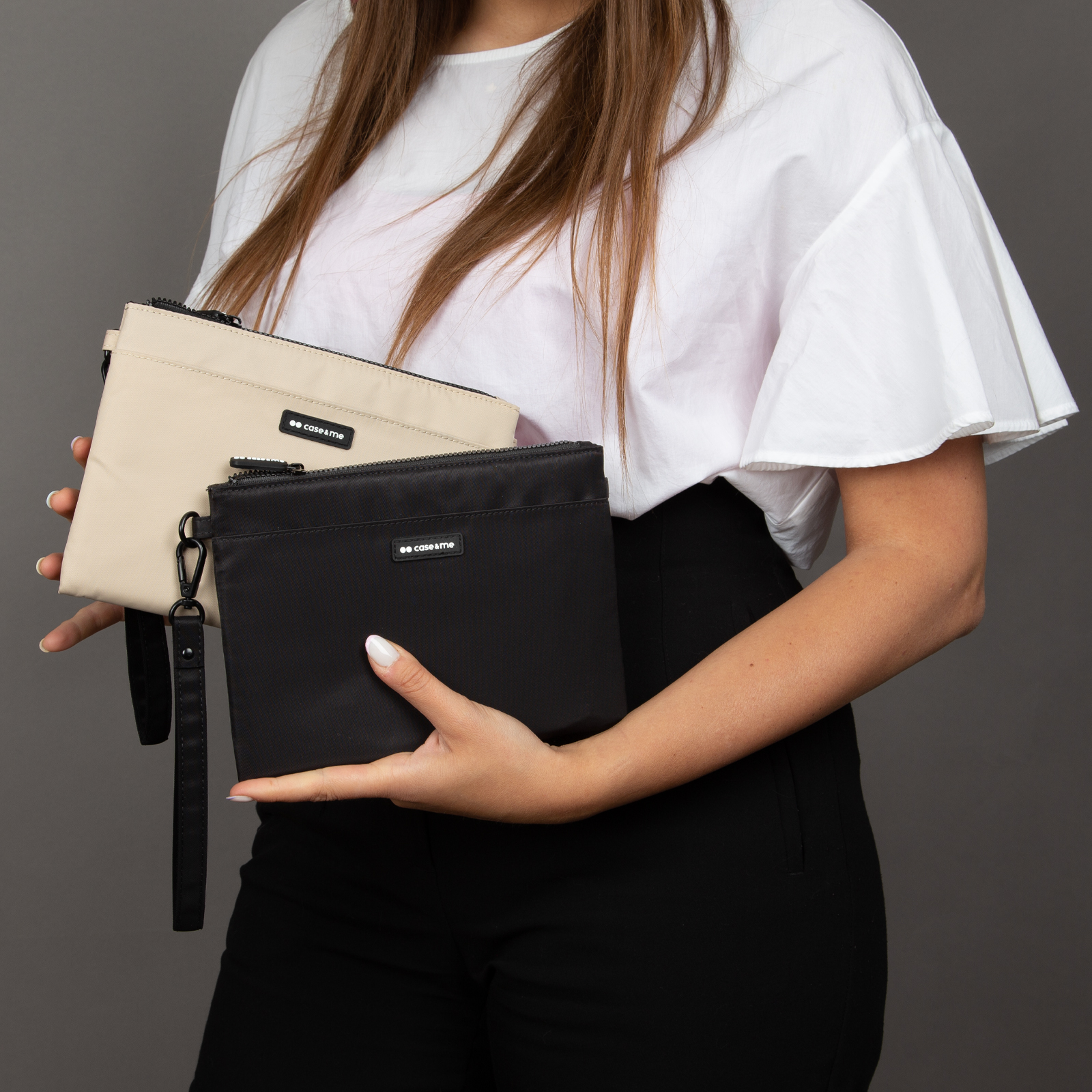 Una mujer con top blanco y pantalón negro sostiene un estuche beige y otro negro con correa de muñeca.