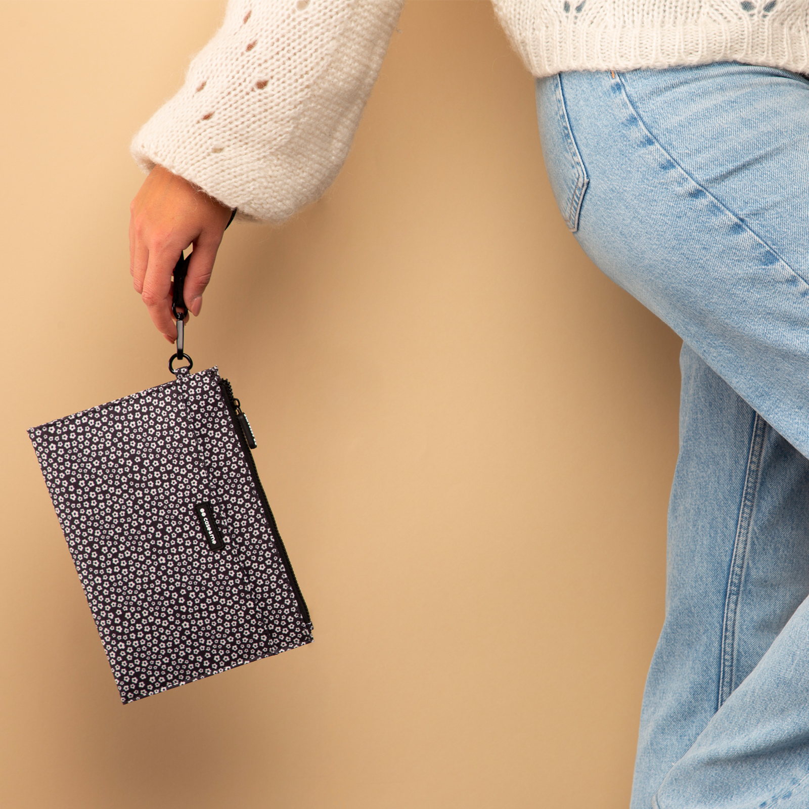 Una persona con suéter blanco y jeans sostiene un pequeño bolso floral negro y blanco sobre fondo beige.