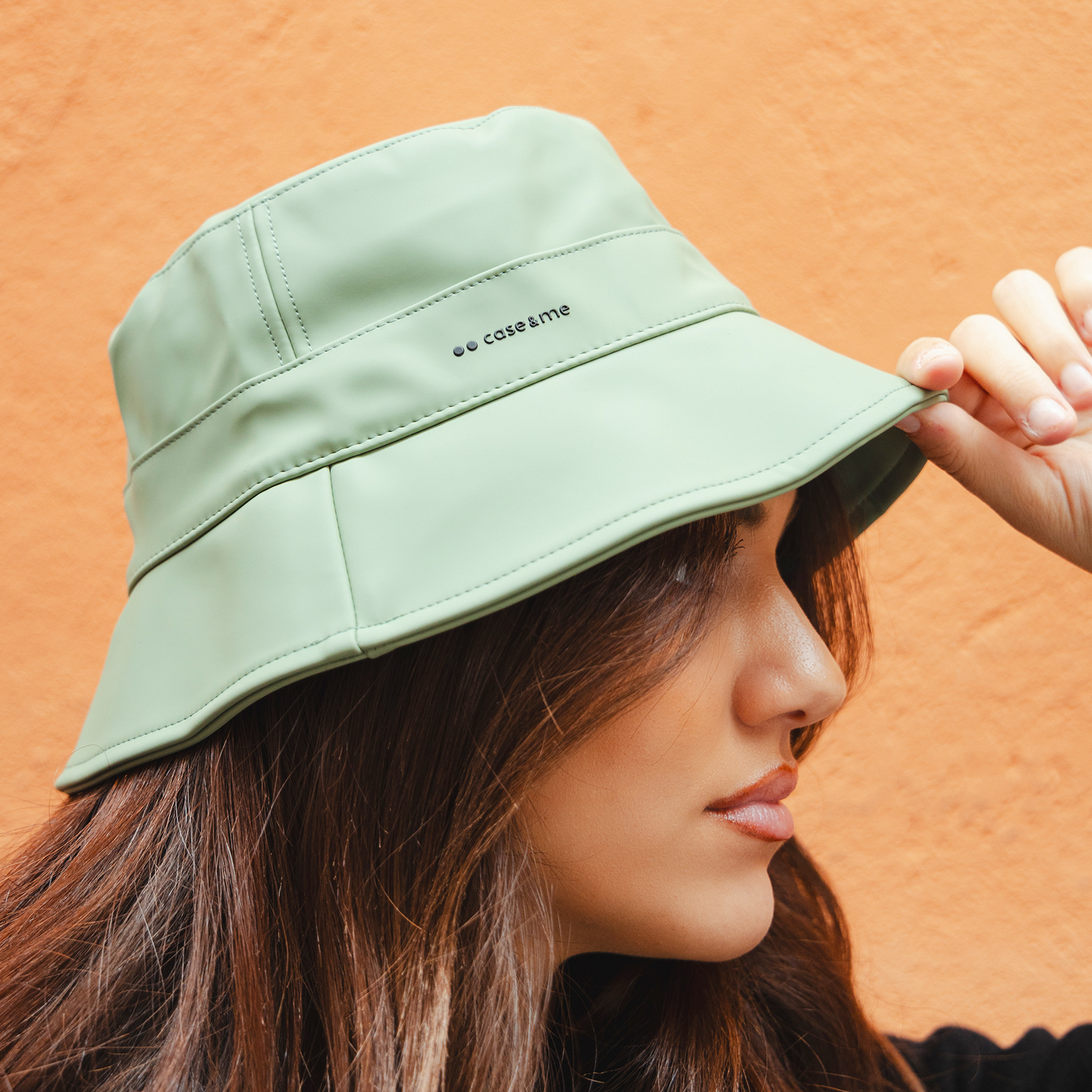Una persona con cabello castaño lleva un sombrero de pescador verde claro contra un fondo naranja.
