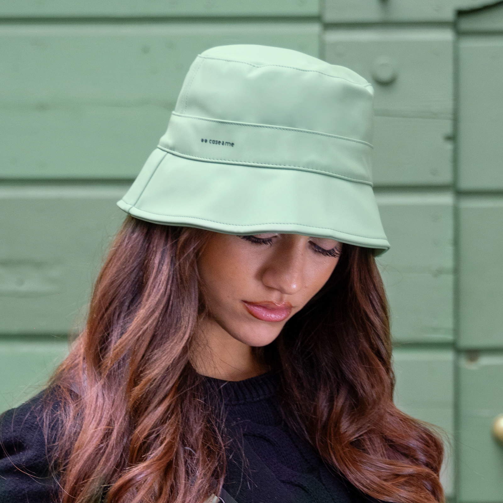 Una mujer con cabello castaño largo lleva un sombrero de pescador verde claro y jersey negro.