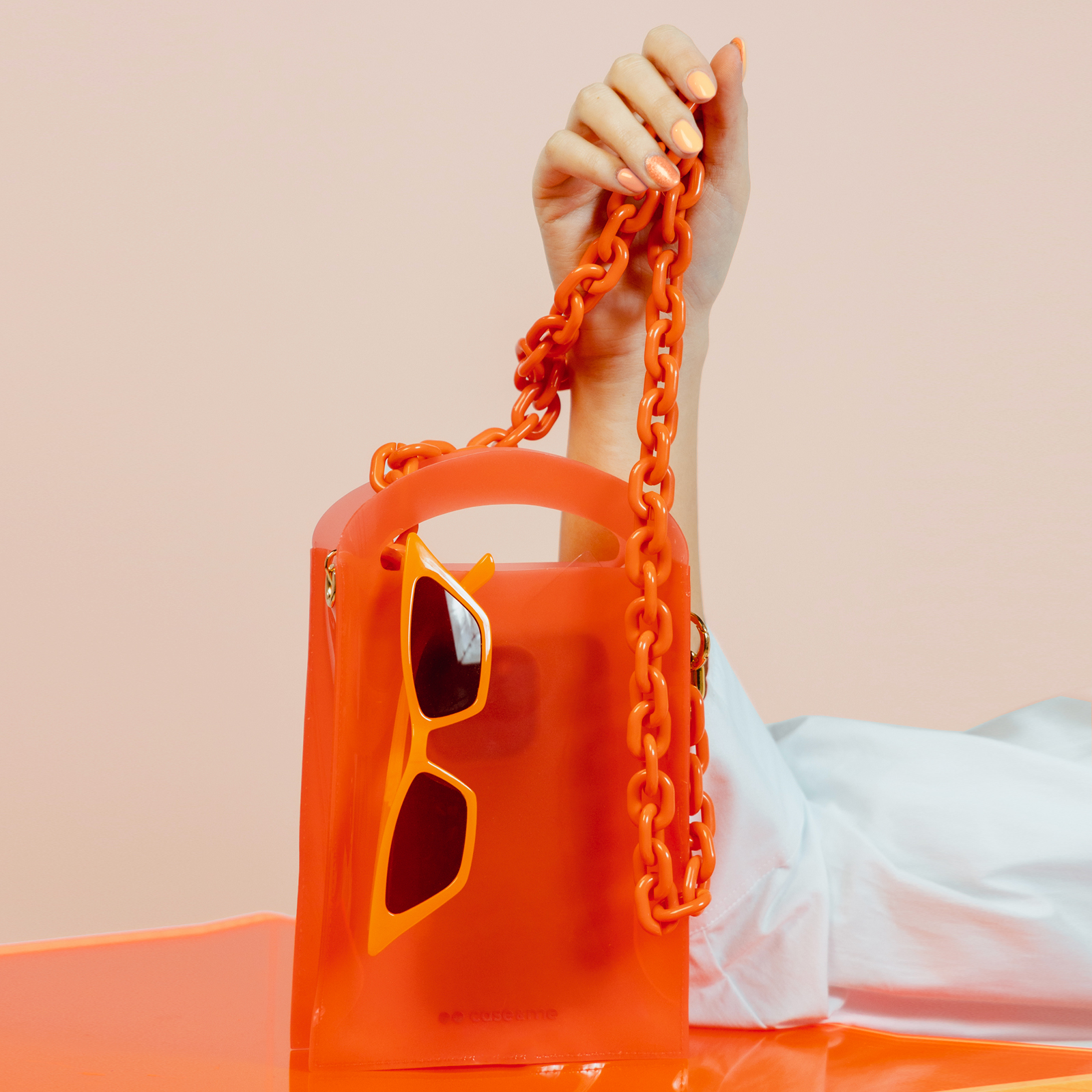 Una mano sostiene una bolsa de plástico naranja con correa de cadena gruesa y gafas a juego.