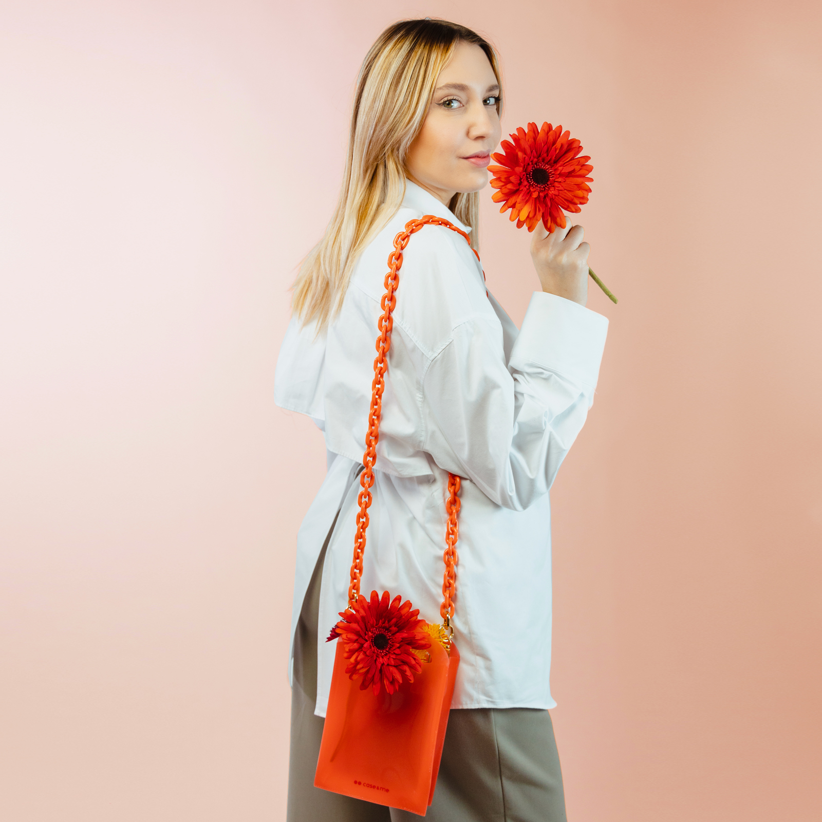 Una mujer rubia con camisa blanca sostiene una flor roja y lleva un bolso naranja sobre fondo rosa.