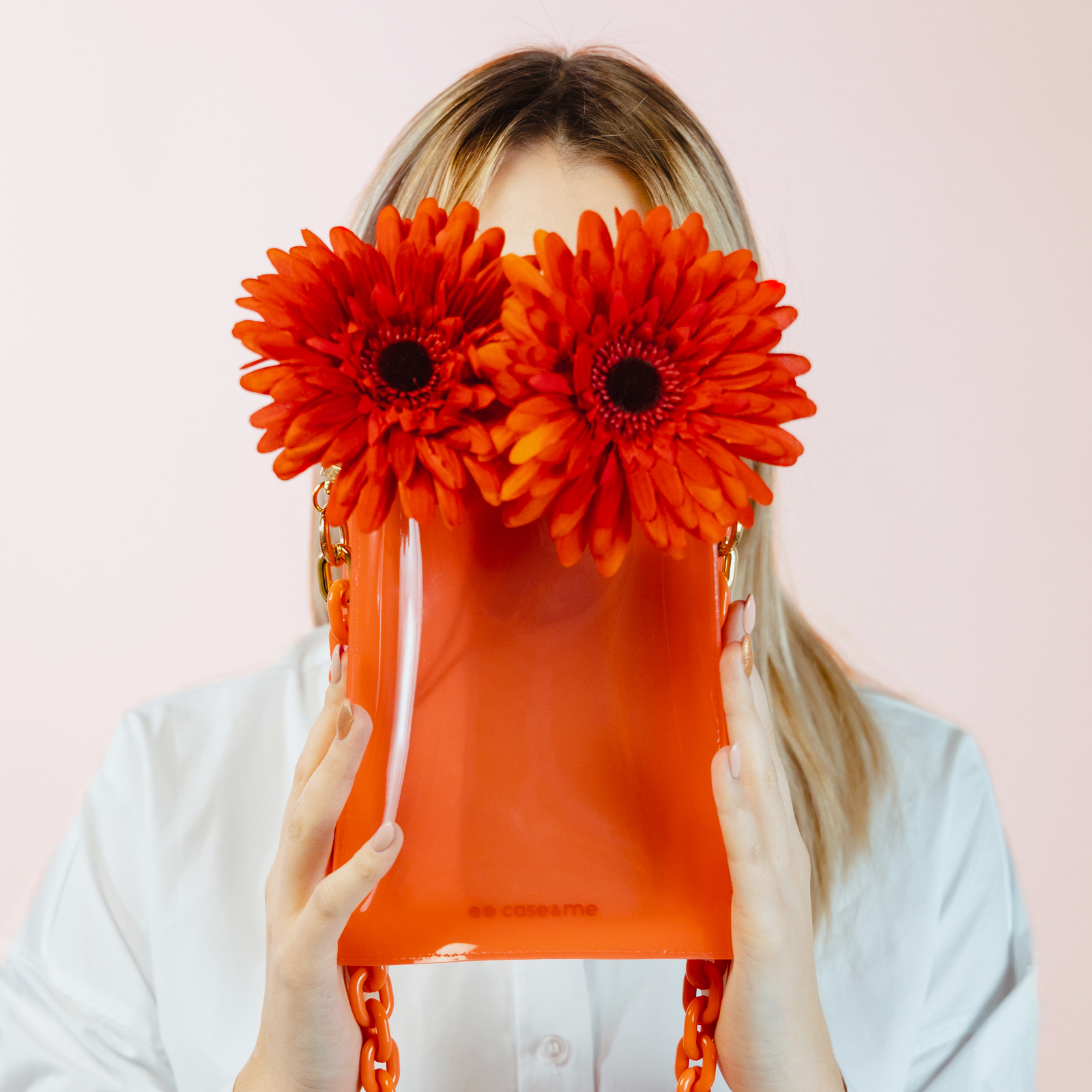 Una mujer rubia con camisa blanca sostiene un bolso naranja con dos grandes flores naranjas cubriendo su cara ante un fondo rosa.