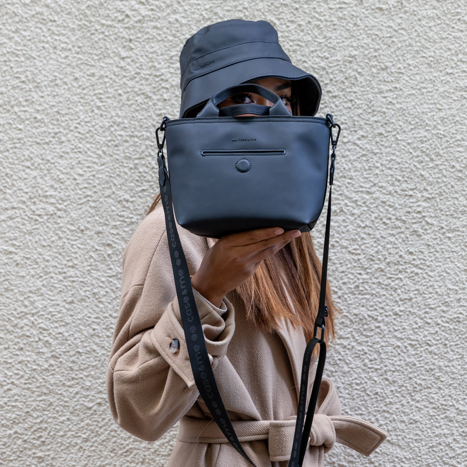 Una mujer con abrigo beige y sombrero negro sostiene un bolso negro frente a su cara.