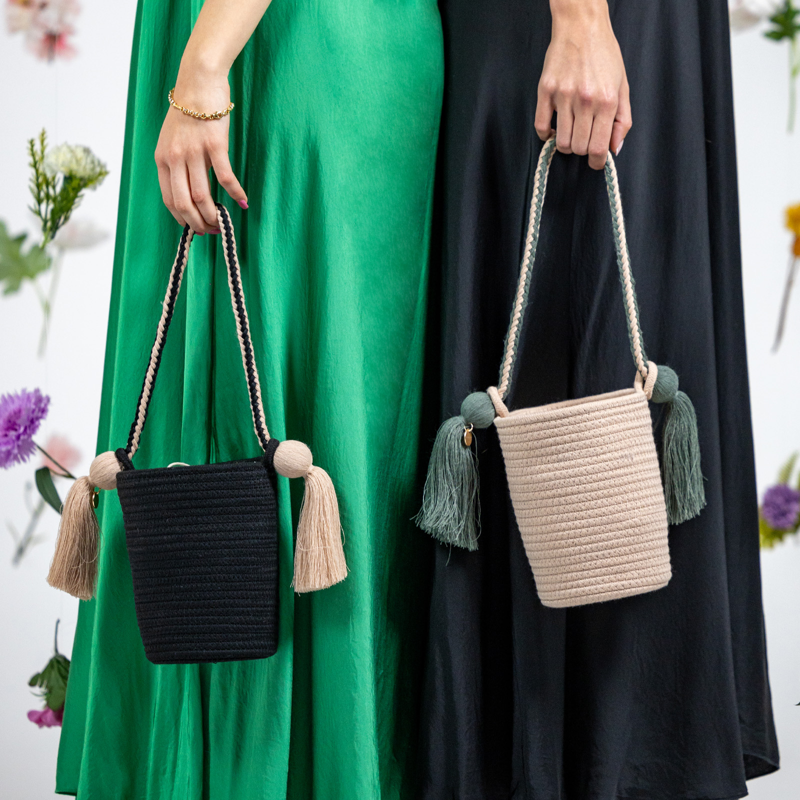 Dos mujeres con vestidos verde y negro sostienen bolsos trenzados con borlas ante un fondo de flores blancas.