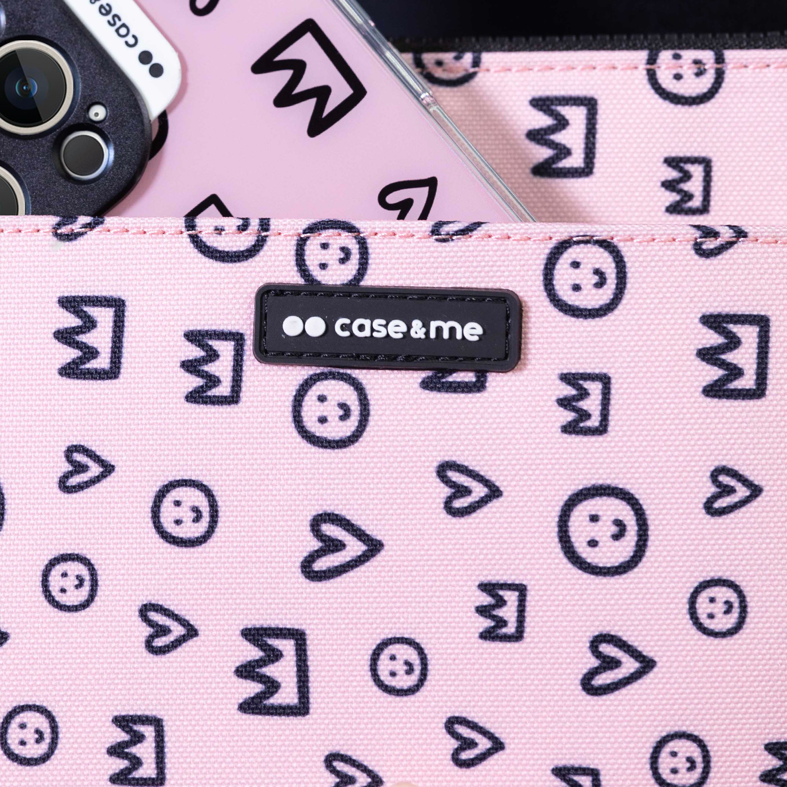Bolso de tela rosa con caras sonrientes, corazones y coronas negras. Un smartphone con funda case&me asoma del bolsillo.