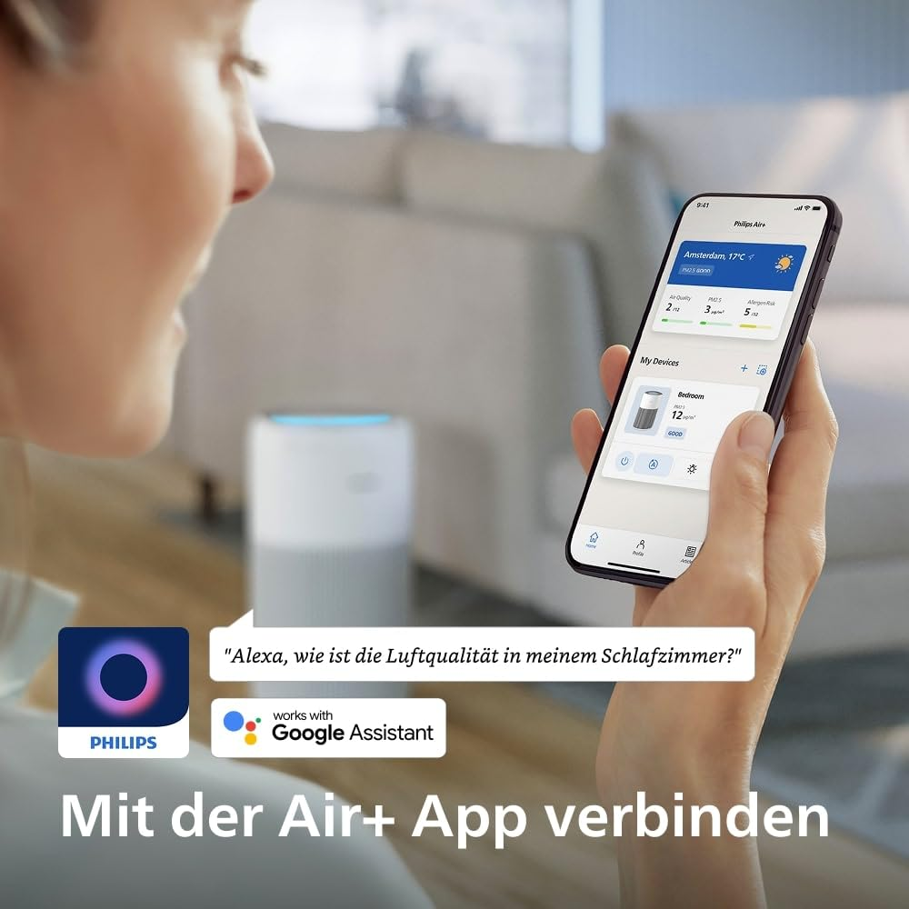Eine Frau hält ein Smartphone mit der Air+ App und spricht mit einem Sprachassistenten, im Hintergrund ein Luftreiniger.