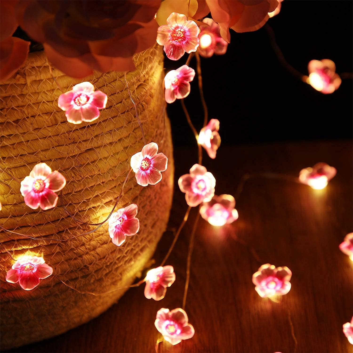 Lichterkette mit rosa, blütenförmigen LED-Lichtern um eine gewebte Vase auf einem Holztisch.
