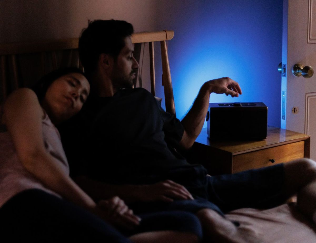Ein Mann sitzt im Bett neben einer schlafenden Frau und greift im gedimmten blauen Licht nach einem Lautsprecher.
