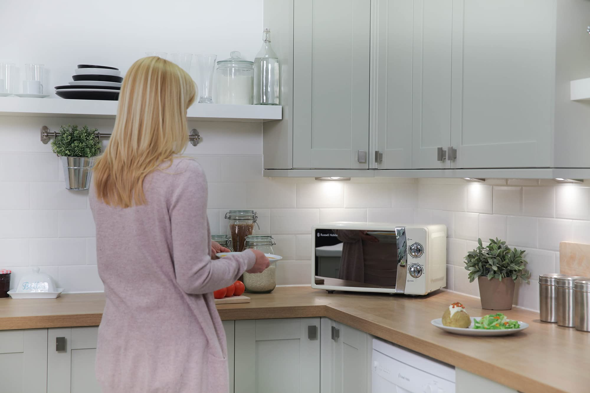 Una donna bionda con giacca rosa in una cucina luminosa di fronte a un microonde crema.