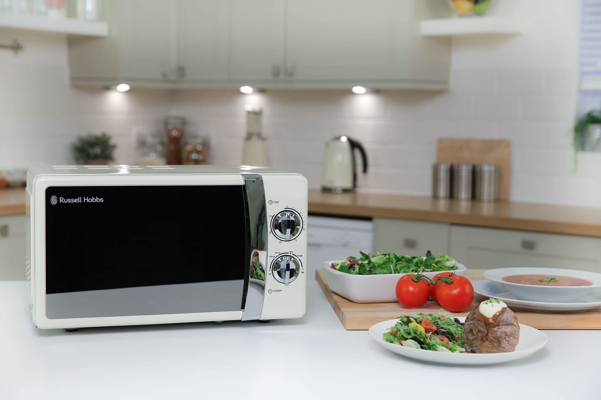Un microonde color crema Russell Hobbs su un bancone bianco in una cucina luminosa, vicino a insalate e patate.