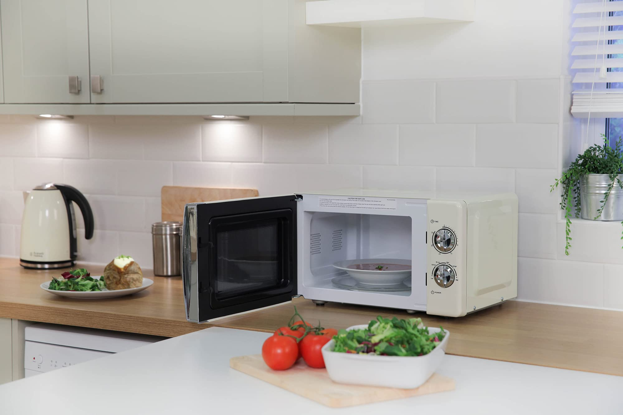 Un microonde beige con la porta aperta su un piano di lavoro in legno in una cucina con insalata e pomodori.