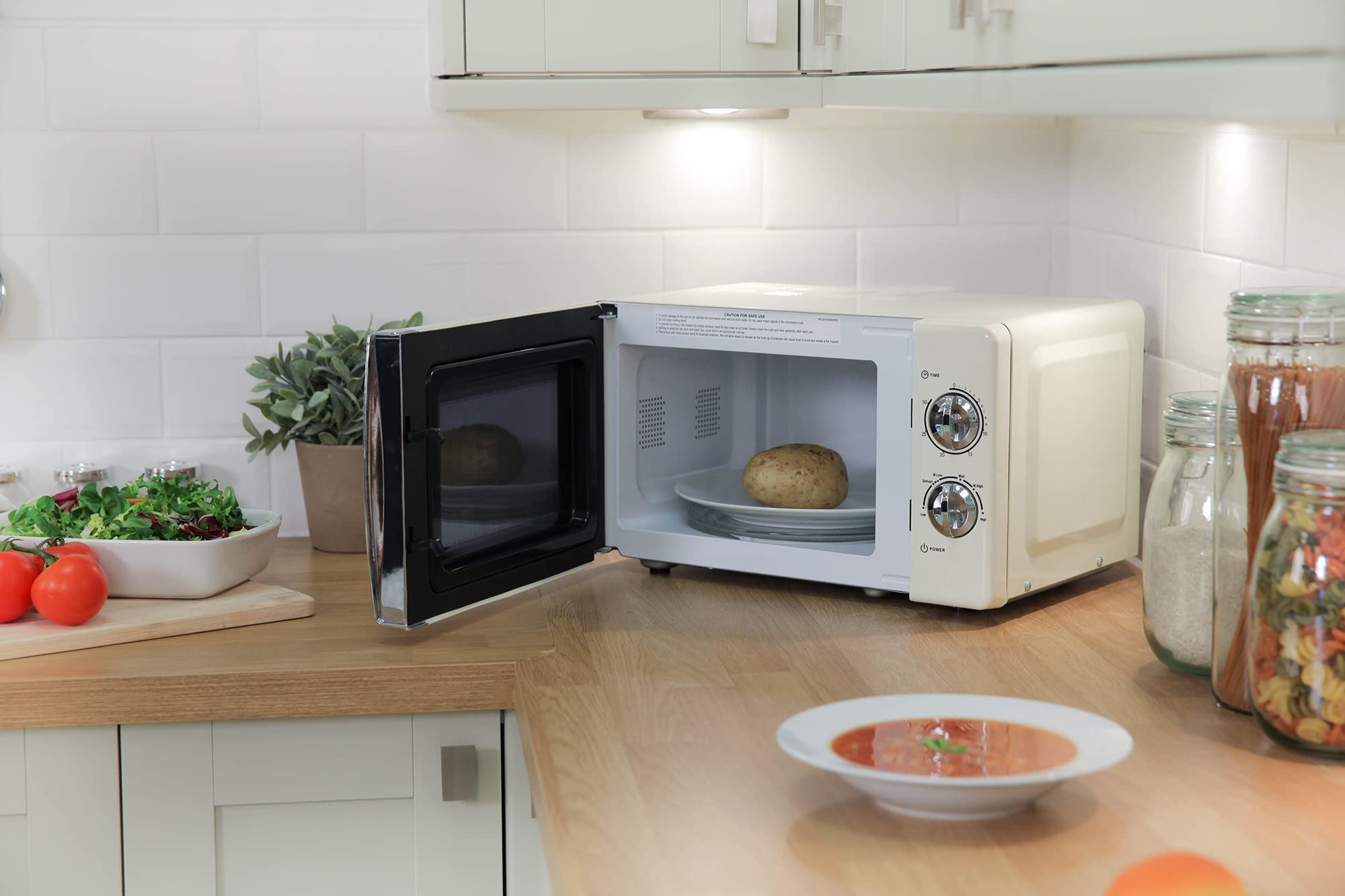 Microonde beige con porta aperta su piano in legno con patata, accanto a insalata, zuppa e barattoli.