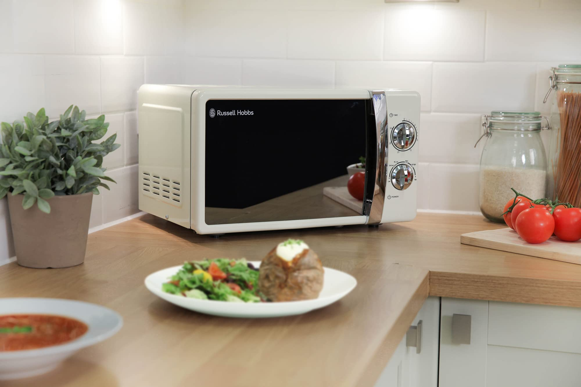 Un forno a microonde Russell Hobbs crema su un piano in legno con insalata, patata al forno e pomodori freschi.