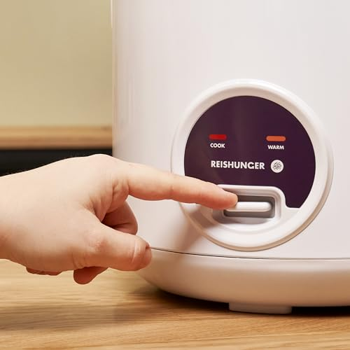 Eine Hand drückt den Schalter eines weißen Reiskochers mit lilafarbenem Bedienfeld auf einem Holztisch.