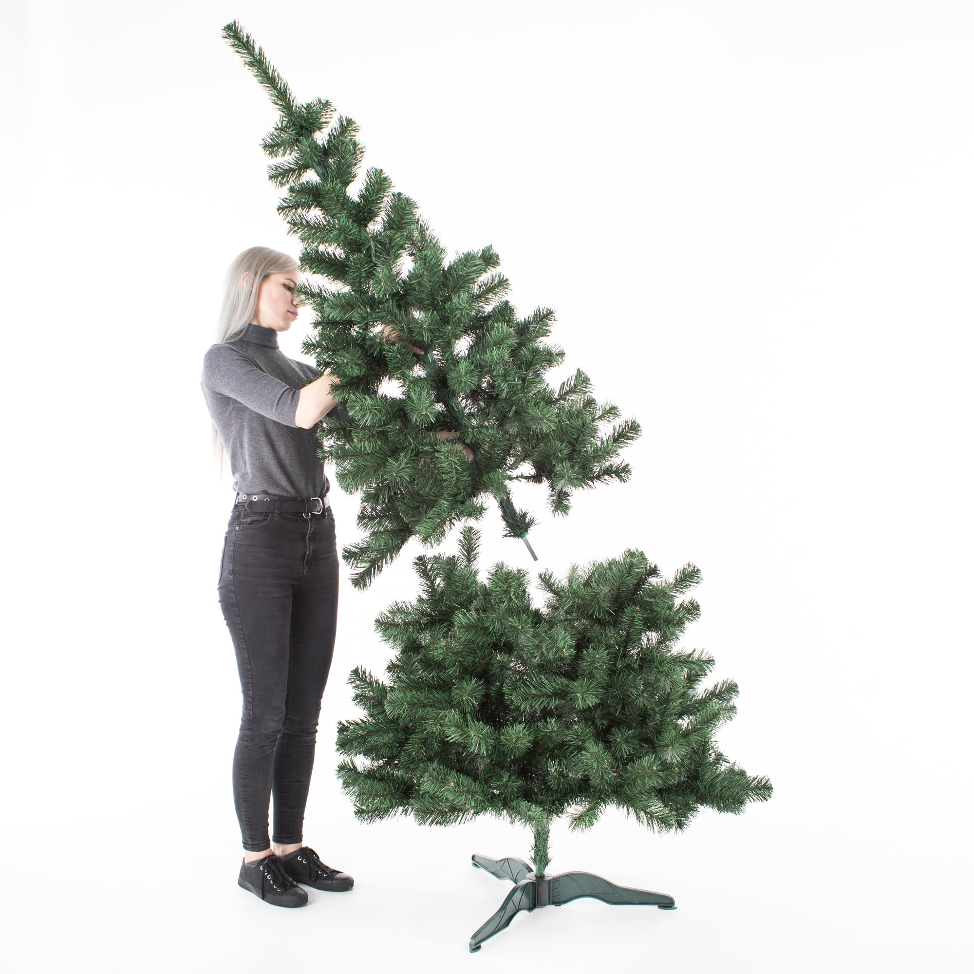 Una donna con maglione grigio a collo alto e jeans neri monta un albero di Natale artificiale.