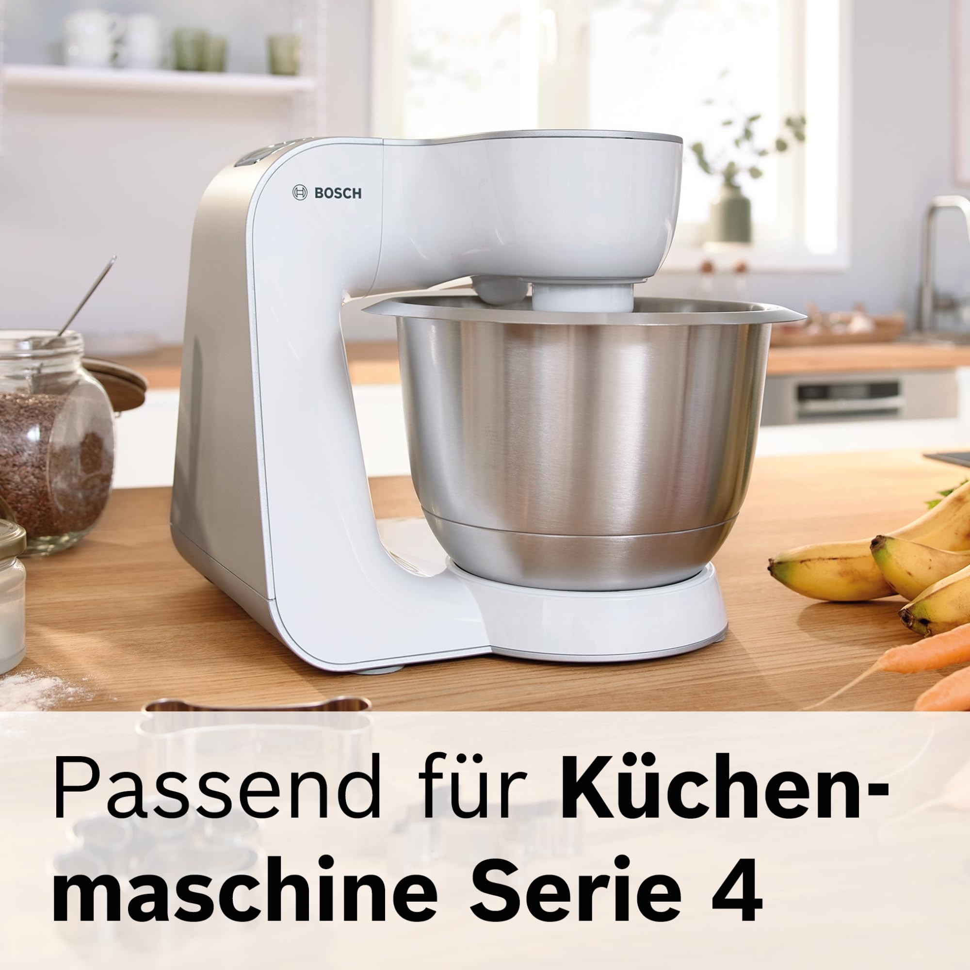 Weiße Bosch Küchenmaschine mit Edelstahlschüssel auf Holzarbeitsplatte, umgeben von Bananen, Karotten und einem Glas.