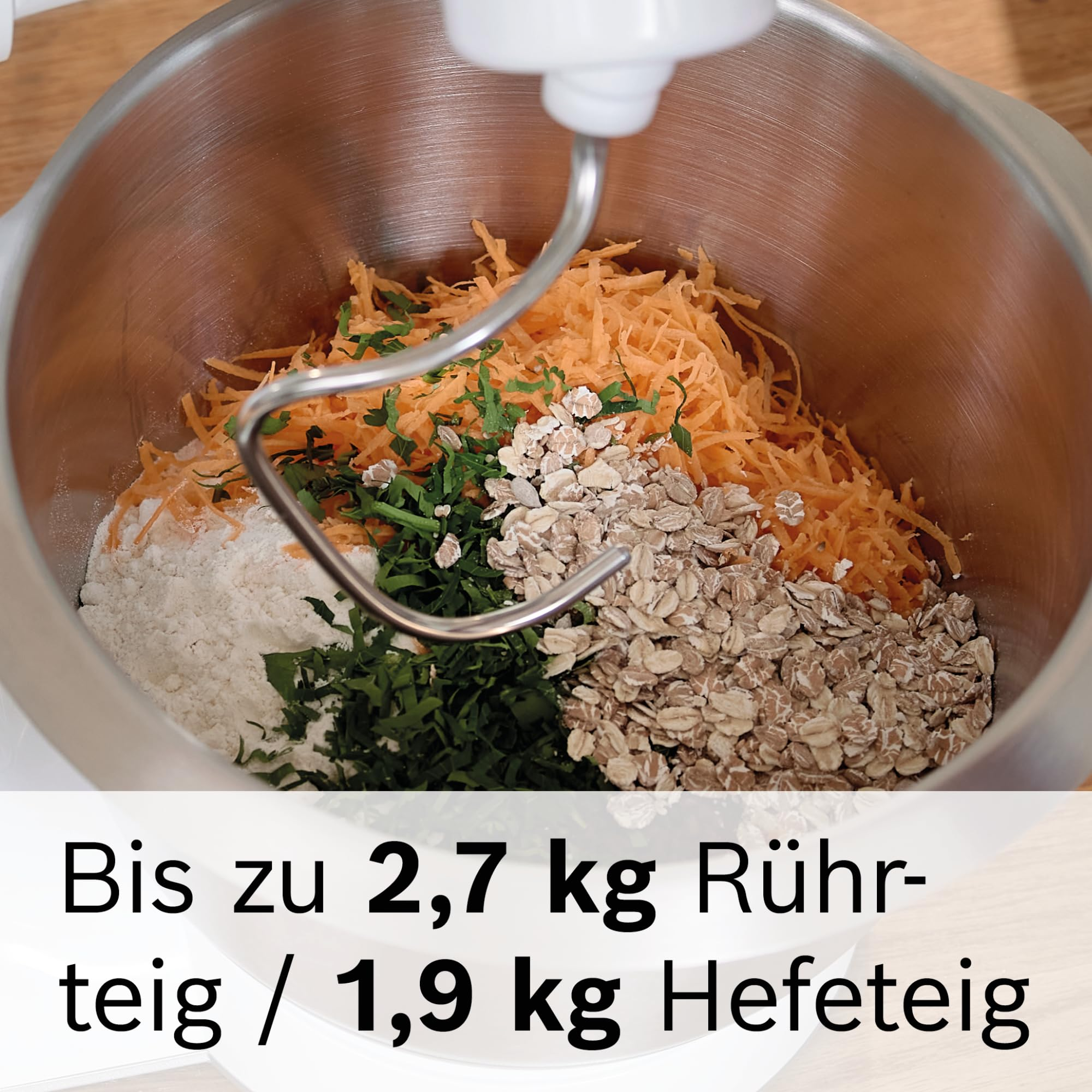 Eine Rührschüssel mit geraspelten Karotten, Haferflocken, Mehl, Kräutern und einem Knethaken.