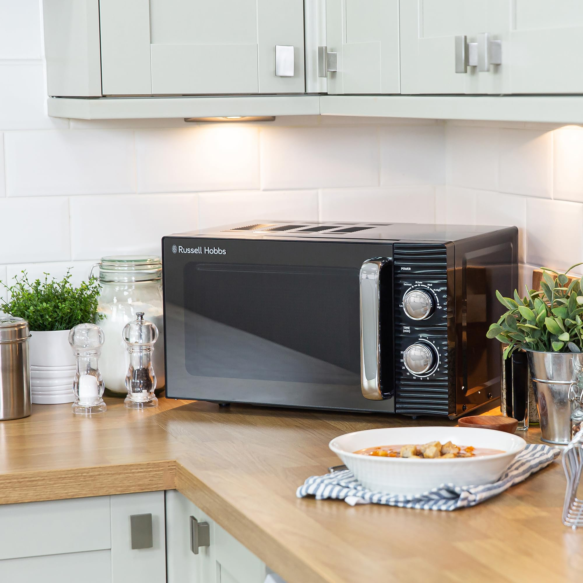 Micro-ondes noir Russell Hobbs sur comptoir en bois avec crédence blanche, épices et plantes.