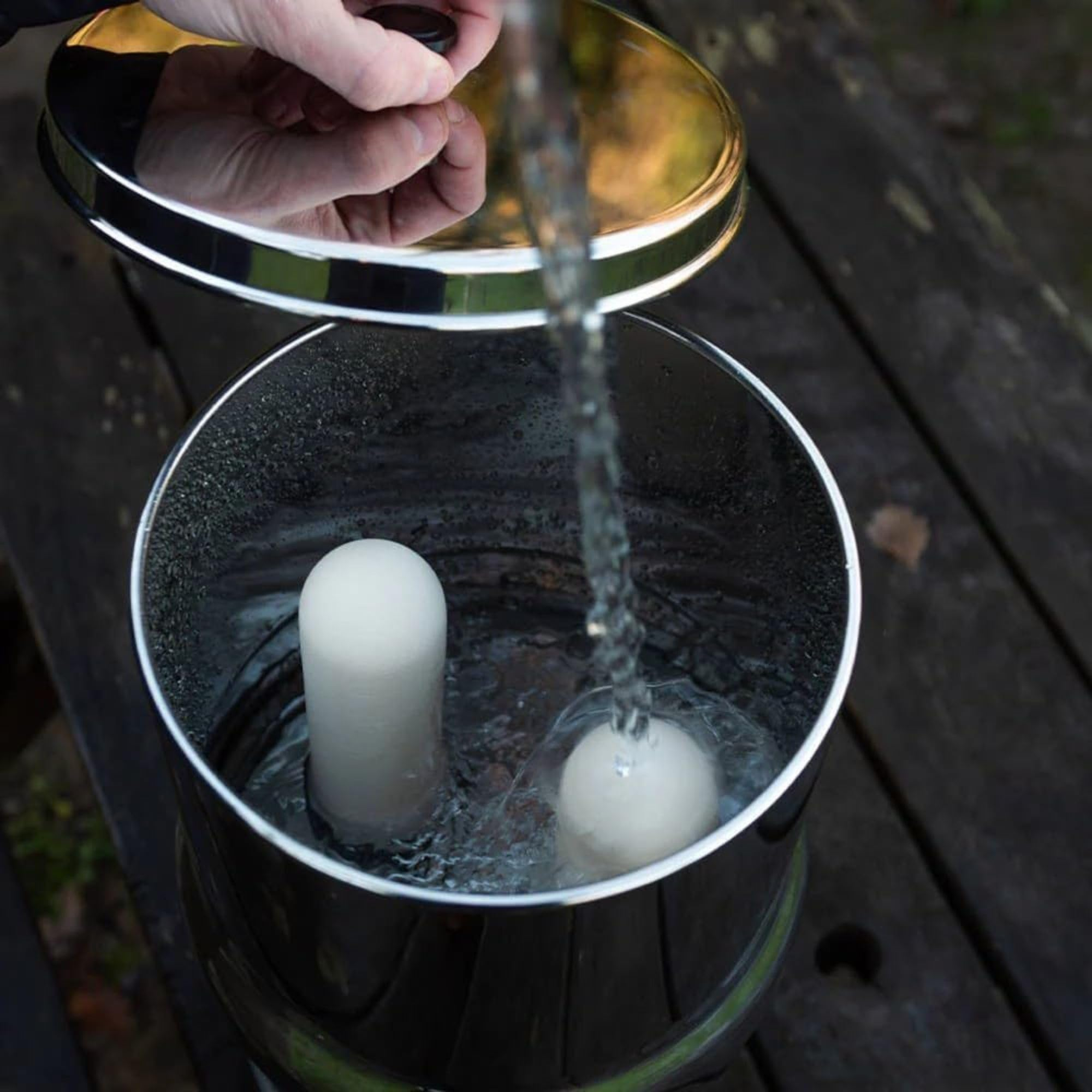 Wasser fließt in einen Edelstahlbehälter mit zwei weißen Keramikfiltern, während eine Hand den Deckel hält.