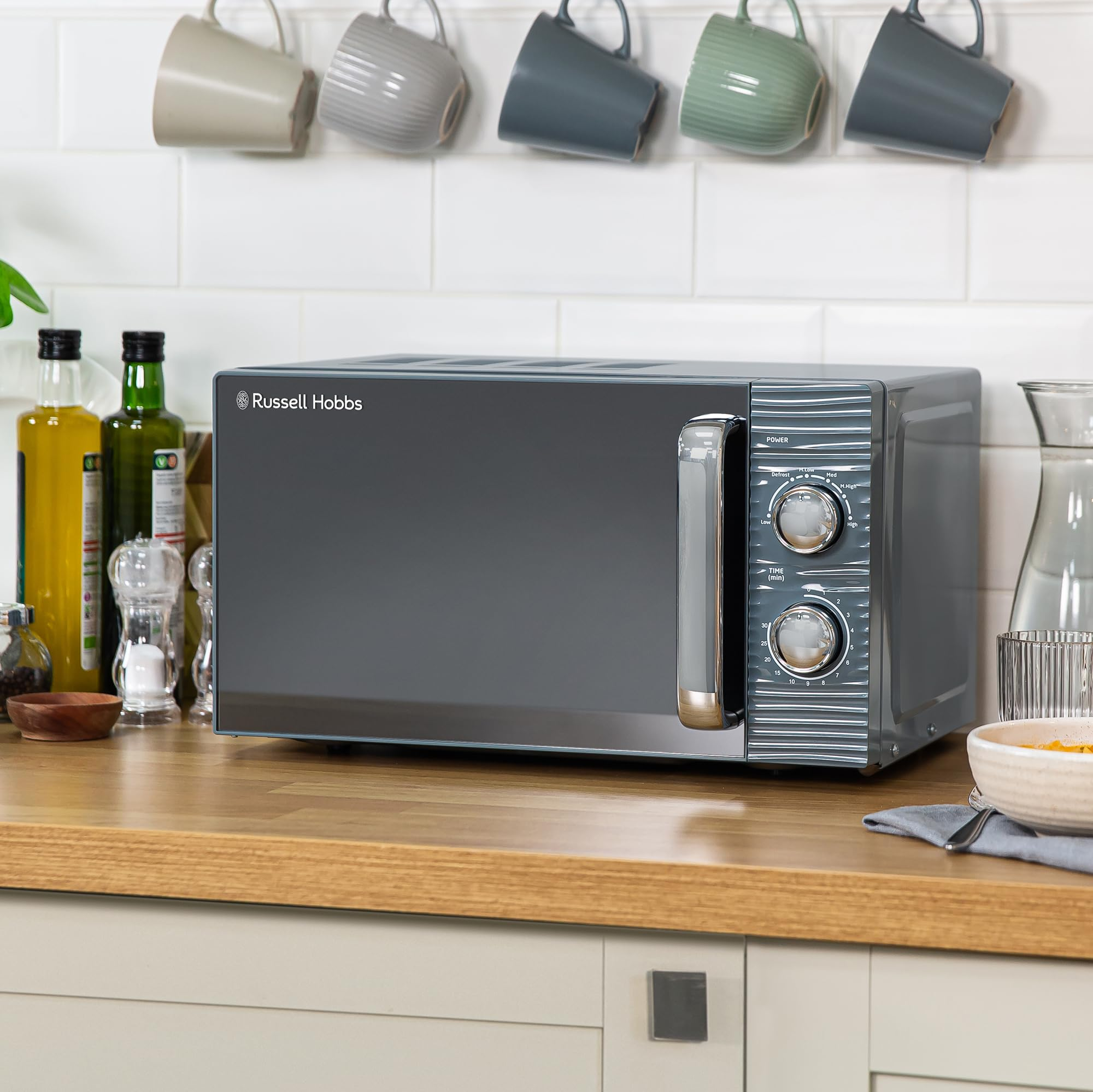 Un micro-ondes gris Russell Hobbs sur un comptoir en bois avec des tasses suspendues et du carrelage blanc.